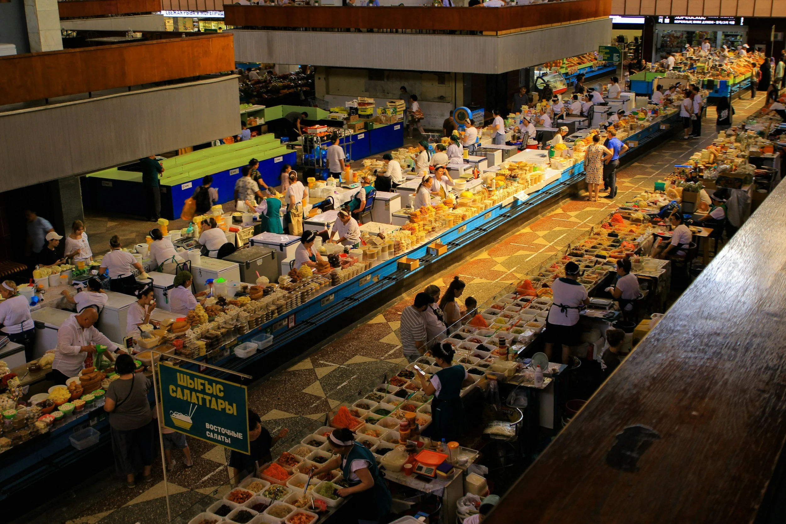 Varios puestos de comercio en un mercado, con vendedores y compradores en un espacio cubierto, con alimentos y productos diversos en mostradores largos.