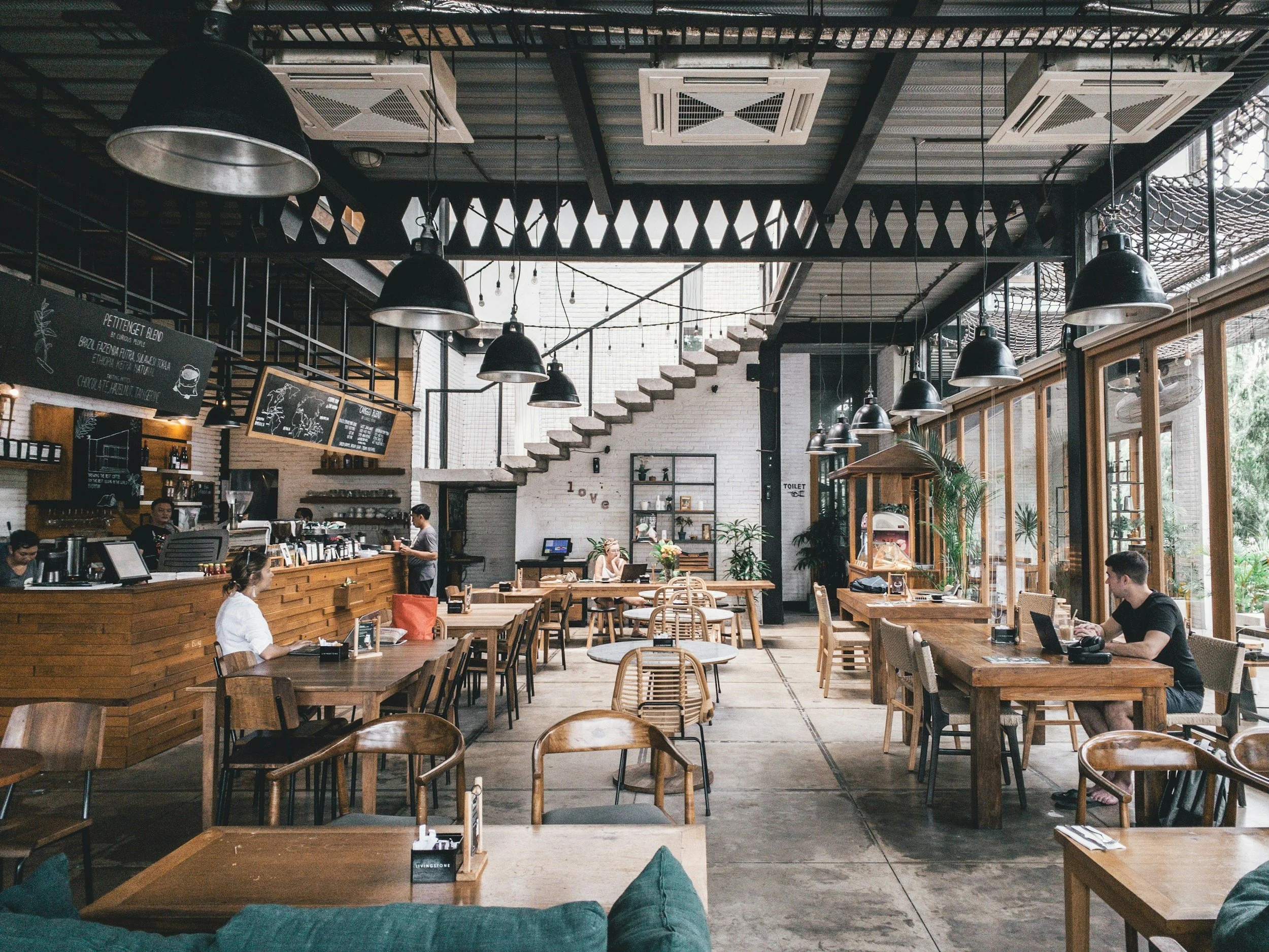 Interior de una cafetería moderna con mesas de madera, sillas y grandes ventanales que dejan entrar mucha luz natural. Hay varias personas sentadas, algunas trabajando en laptops, y un espacio al fondo con una escalera y plantas decorativas.