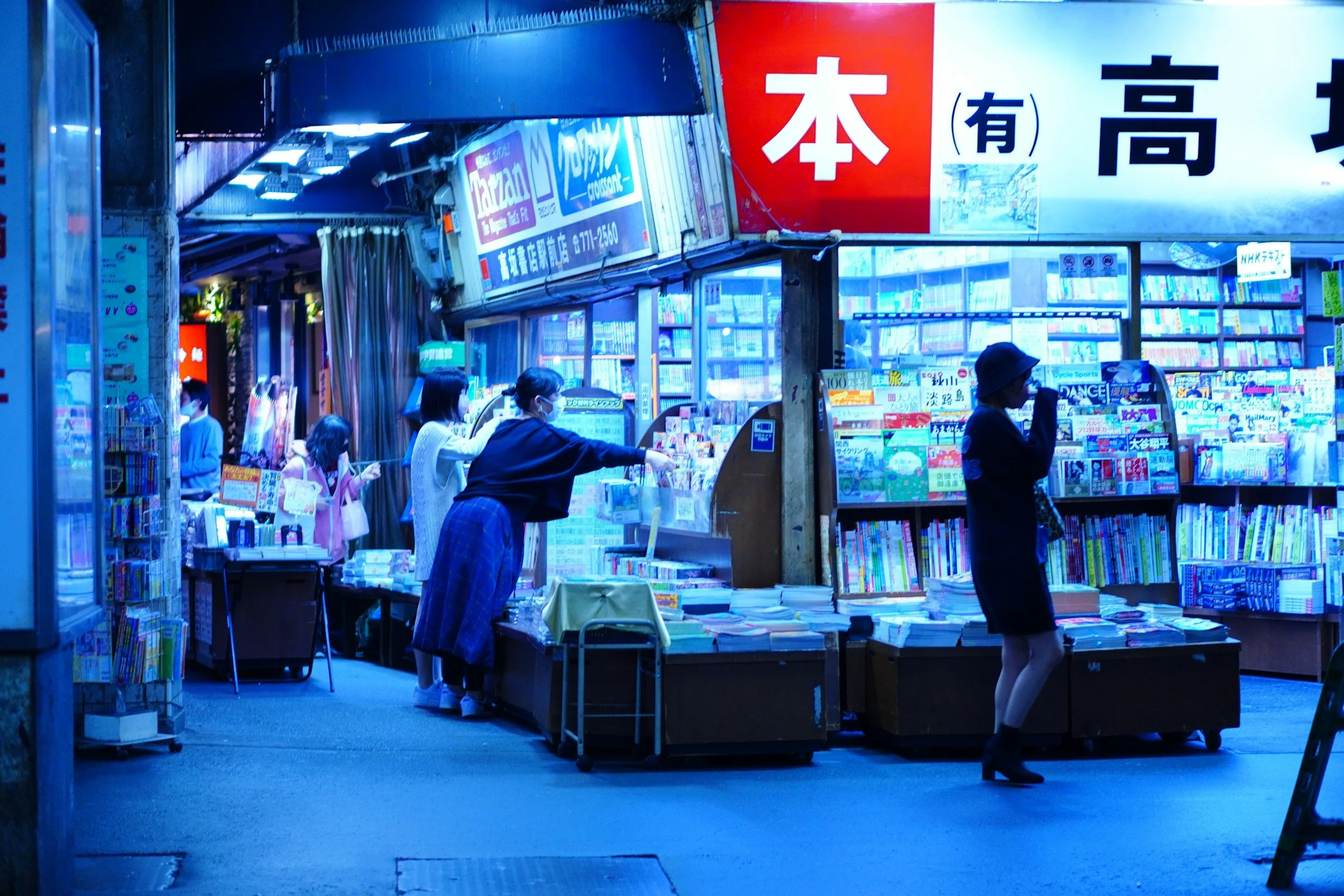 Una tienda de libros en una calle, con varias personas comprando y mirando libros, en un ambiente iluminado con luz azul.