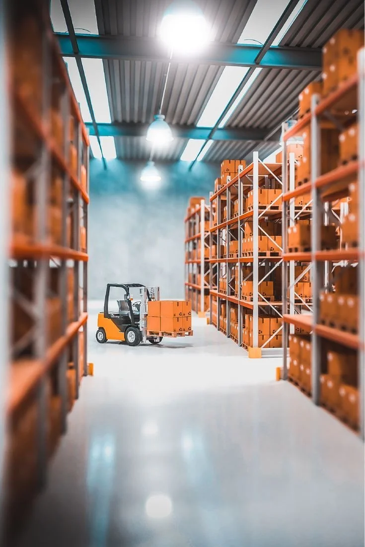Interior de un almacén con estanterías llenas de cajas naranjas y una carretilla elevadora transportando cajas en el pasillo central