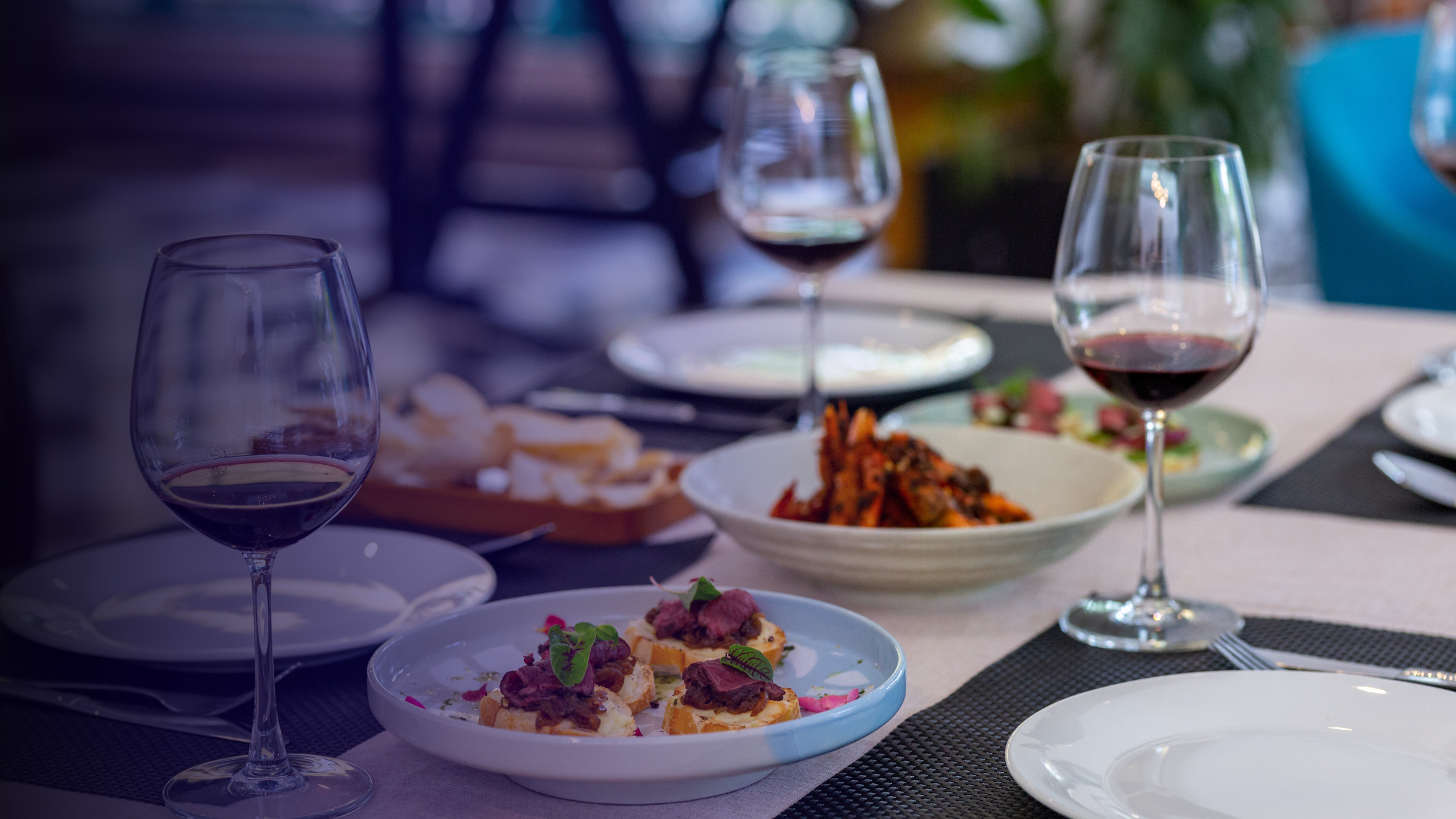 Mesa con copas de vino tinto, varios platos de comida y pequeños bocadillos en un ambiente de restaurante