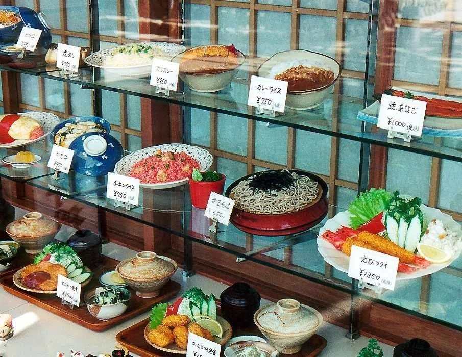 Vitrina de comida japonesa y platos tradicionales, con etiquetas de precios en yenes, incluyendo arroz, fideos, sushi, tempura y otros. Algunos alimentos en platos decorativos y recipientes tradicionales.