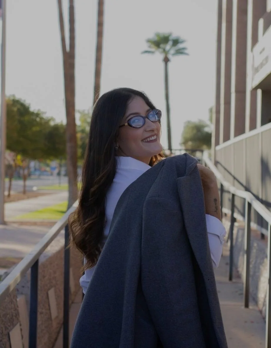Mujer sonriendo con gafas en un entorno exterior, con árboles y edificios en el fondo.