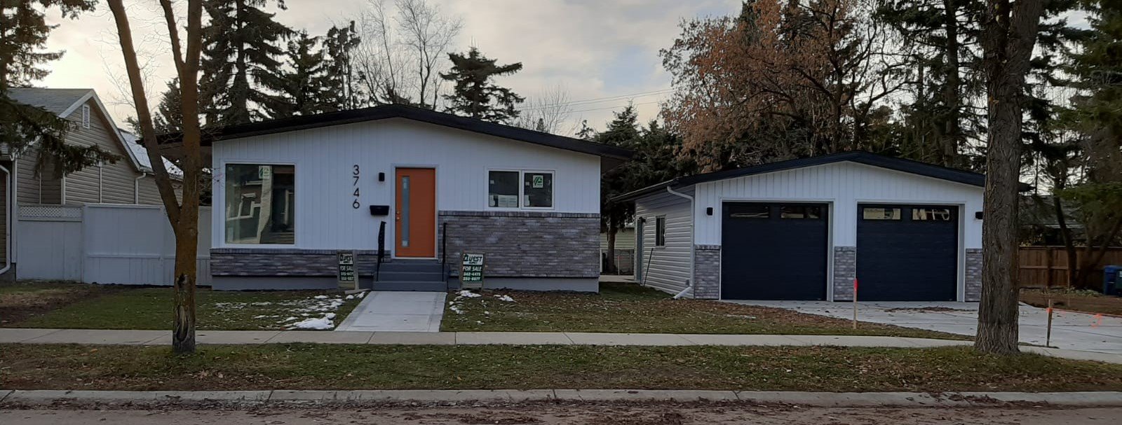 A modern house with a front yard, concrete walkway, and a detached garage, surrounded by trees, on a slightly overcast day.