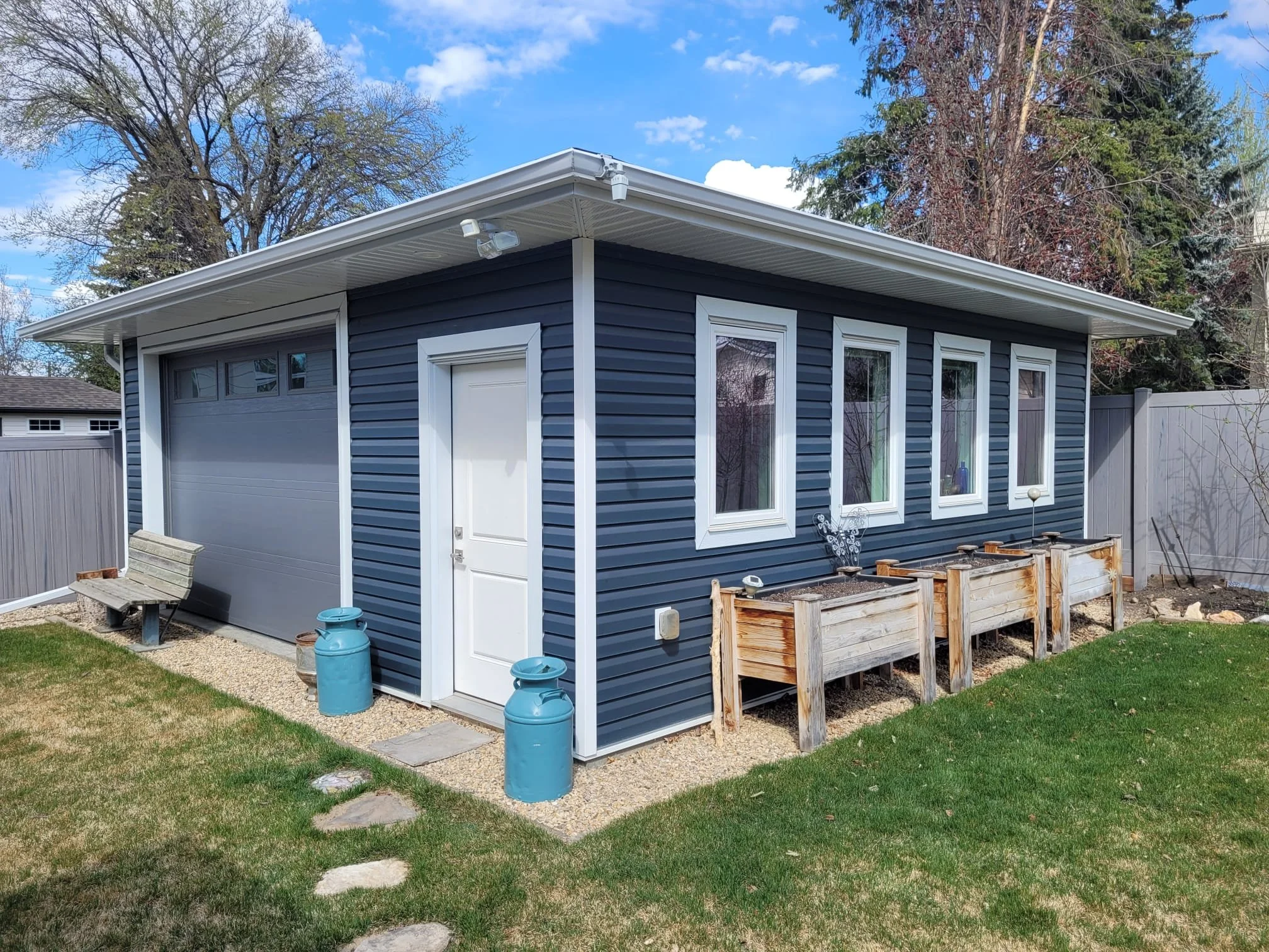 A small blue garage building with white trim, four windows, and a white door in a backyard with a green lawn and a stone pathway. There are two blue milk canisters, a wooden bench, and empty wooden planters outside.