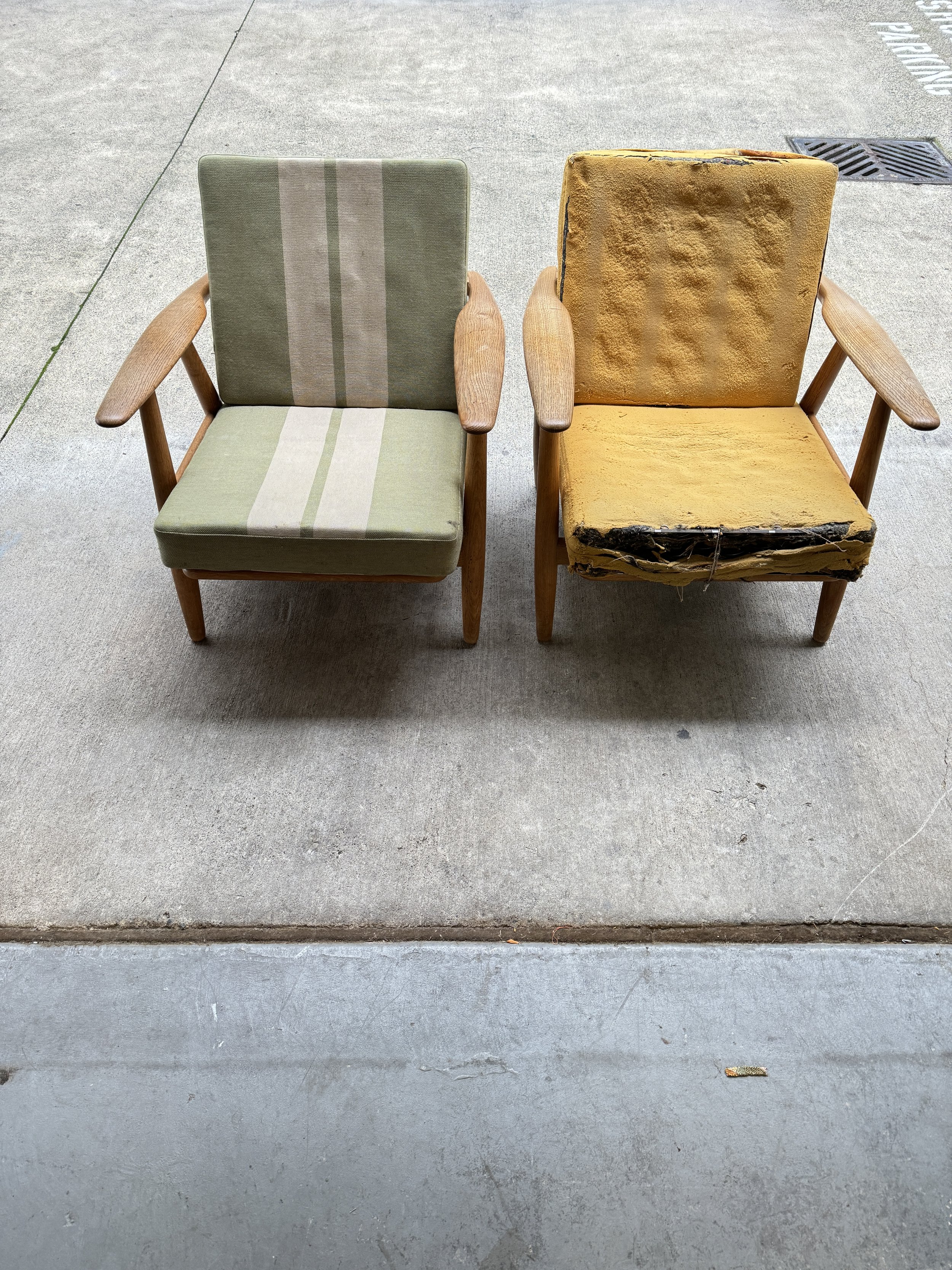 Two vintage mid century modern 'Cigar' armchairs designed by Hans Wegner, prior to restoration, with wooden armrests and legs, one with green and beige striped fabric, the other with yellowed and worn upholstery, on concrete.