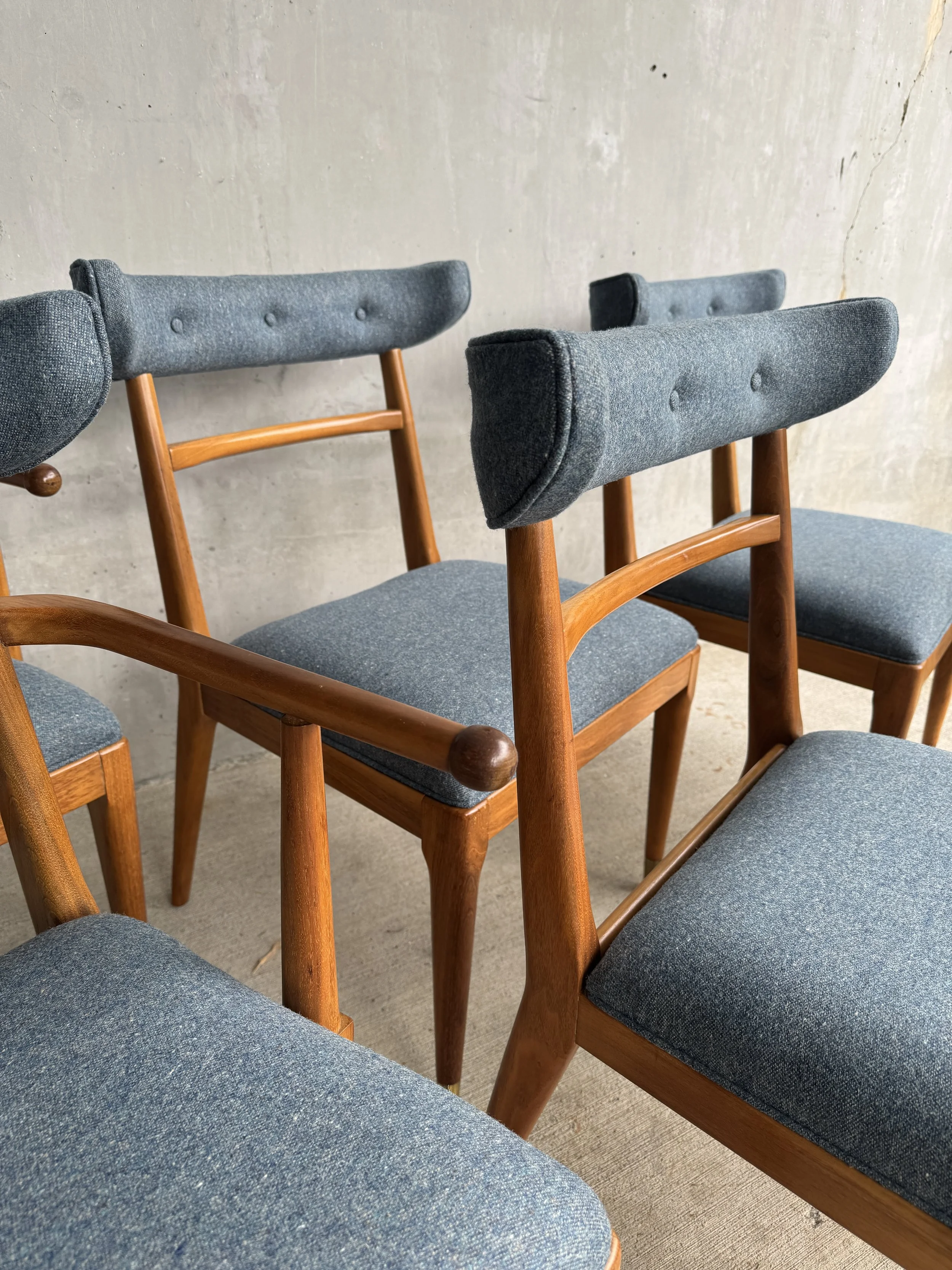 Multiple mid-century modern style chairs with wooden frames and blue upholstered seats and backs arranged against a textured concrete wall.