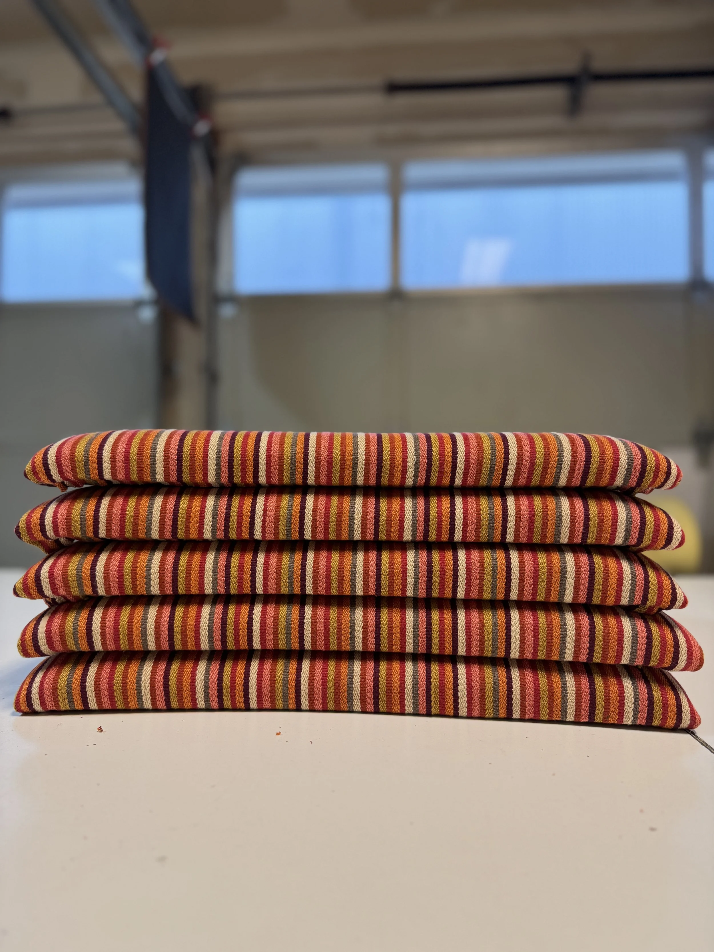 A stack of four colorful, striped cushions on a white surface with a blurred workshop background.