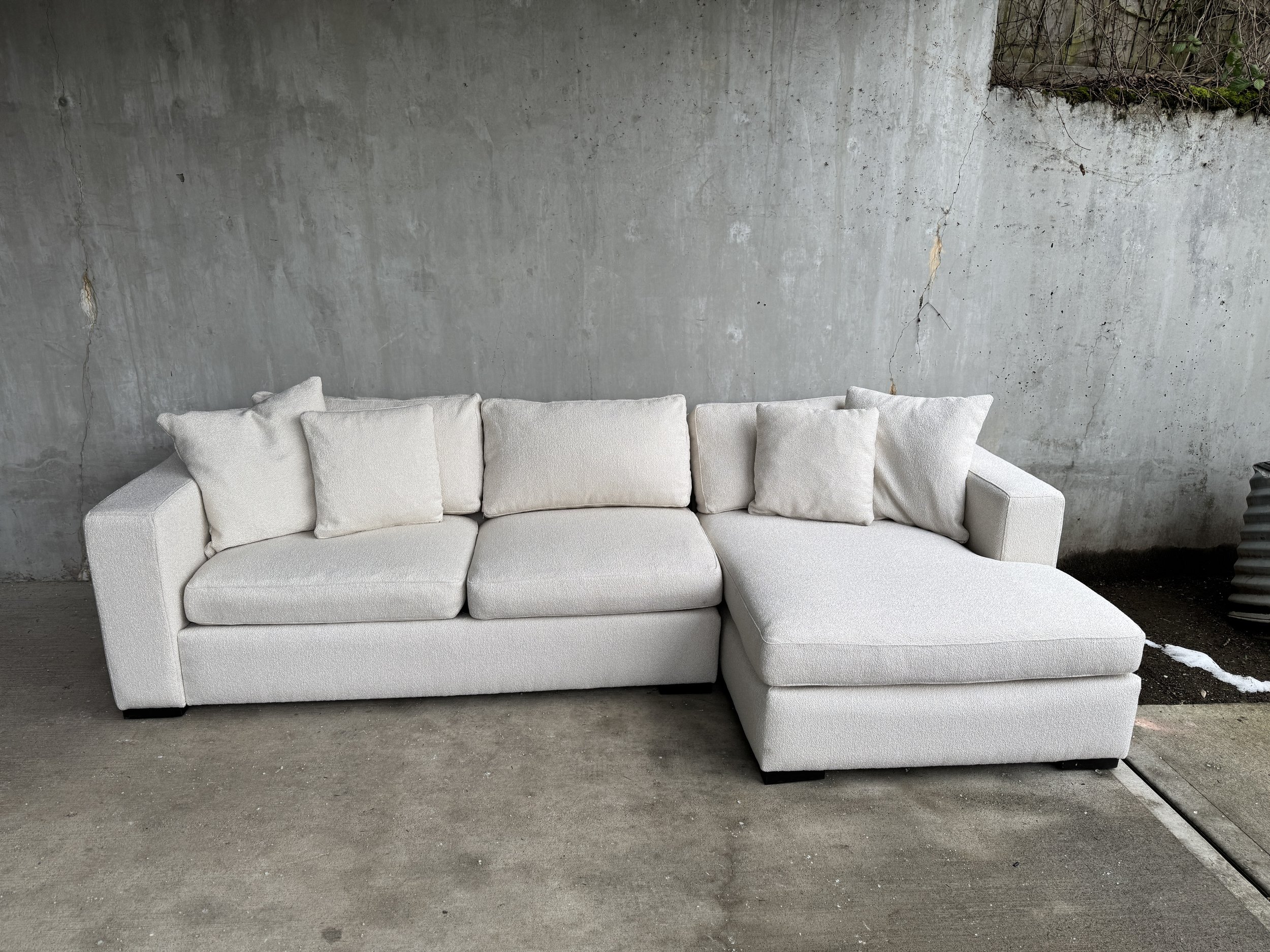A white sectional sofa with multiple cushions sits against a textured concrete wall with cracks and peeling paint. The floor appears to be concrete.