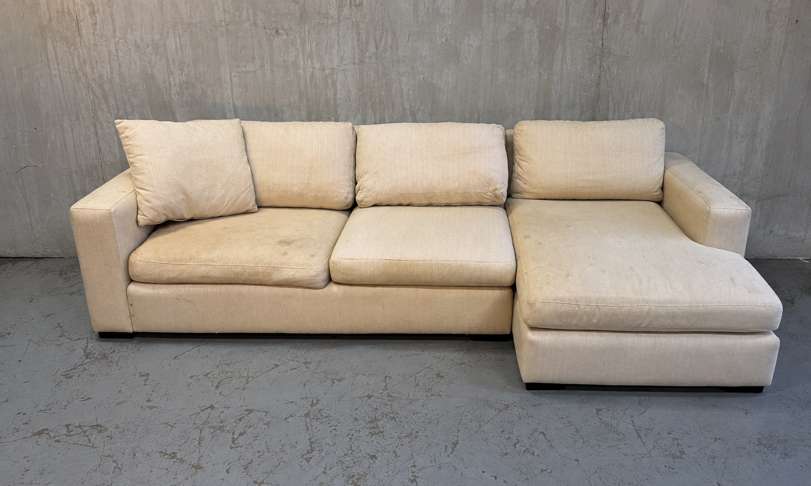 Cream-colored sectional sofa with three cushions and two pillows, placed against a concrete wall in a minimalist space.
