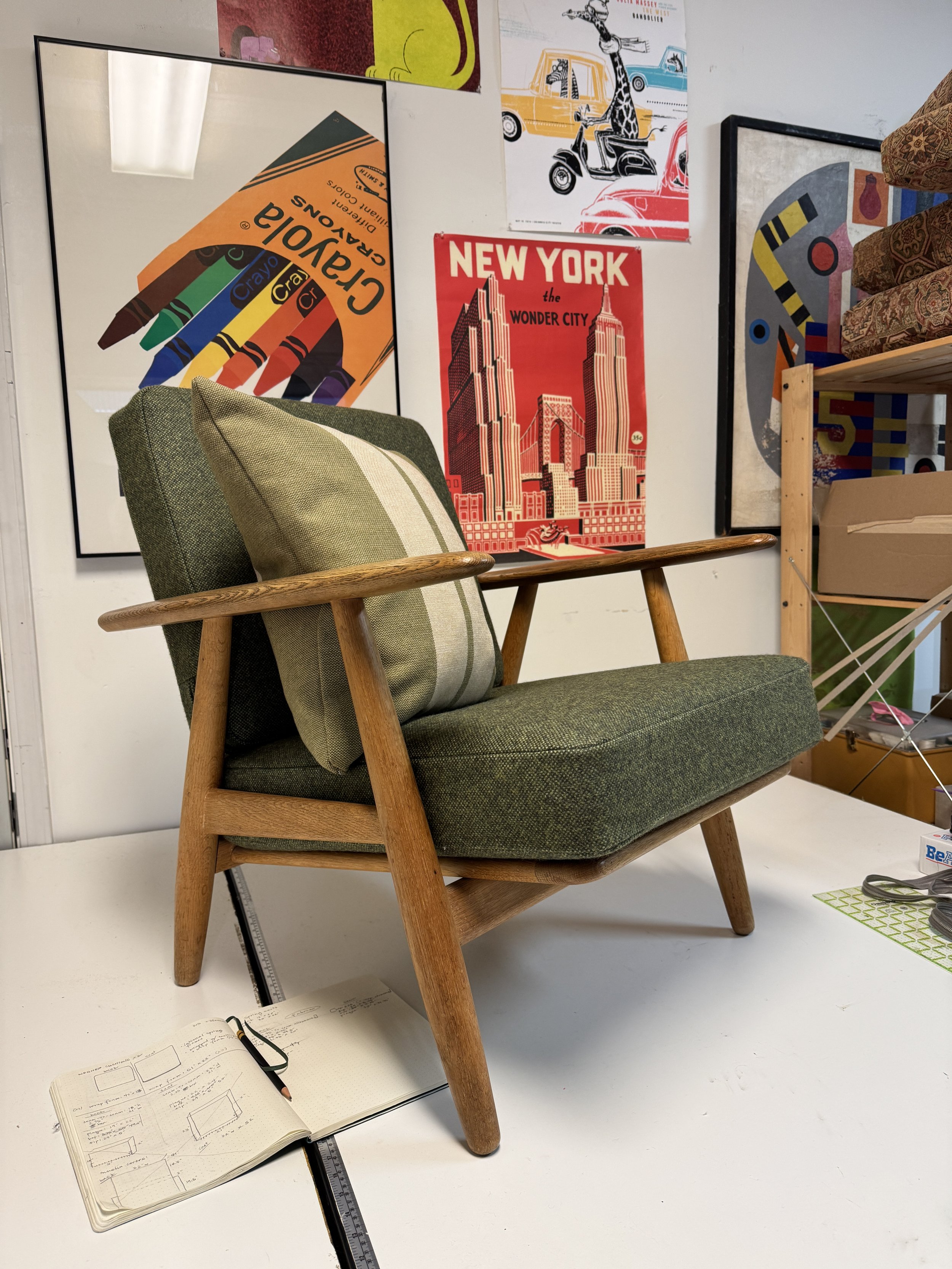 A newly upholstered mid century modern 'Cigar' armchair by Hans Wegner with green cushions and wooden frame on a white surface, with a hardcover open notebook, black pen, and ruler laid across the surface. The background features vintage posters.