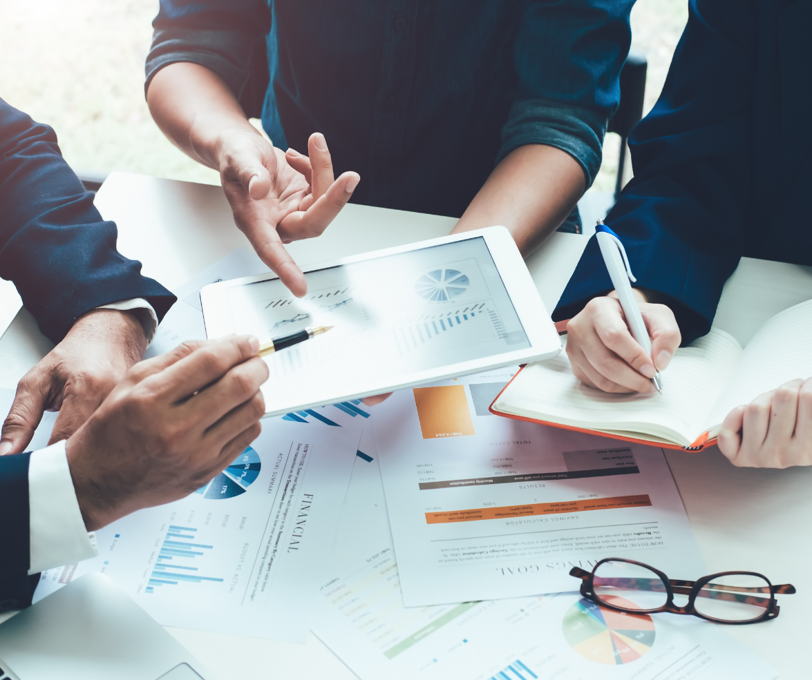 Businesspeople at a meeting reviewing financial charts and graphs on a tablet and paper documents, with glasses and a notebook on the table.