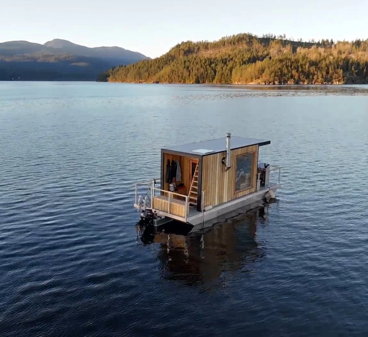 A floating tiny house on a calm lake with forested hills and mountains in the background, featuring wooden exterior walls, a small deck, and outdoor furniture.