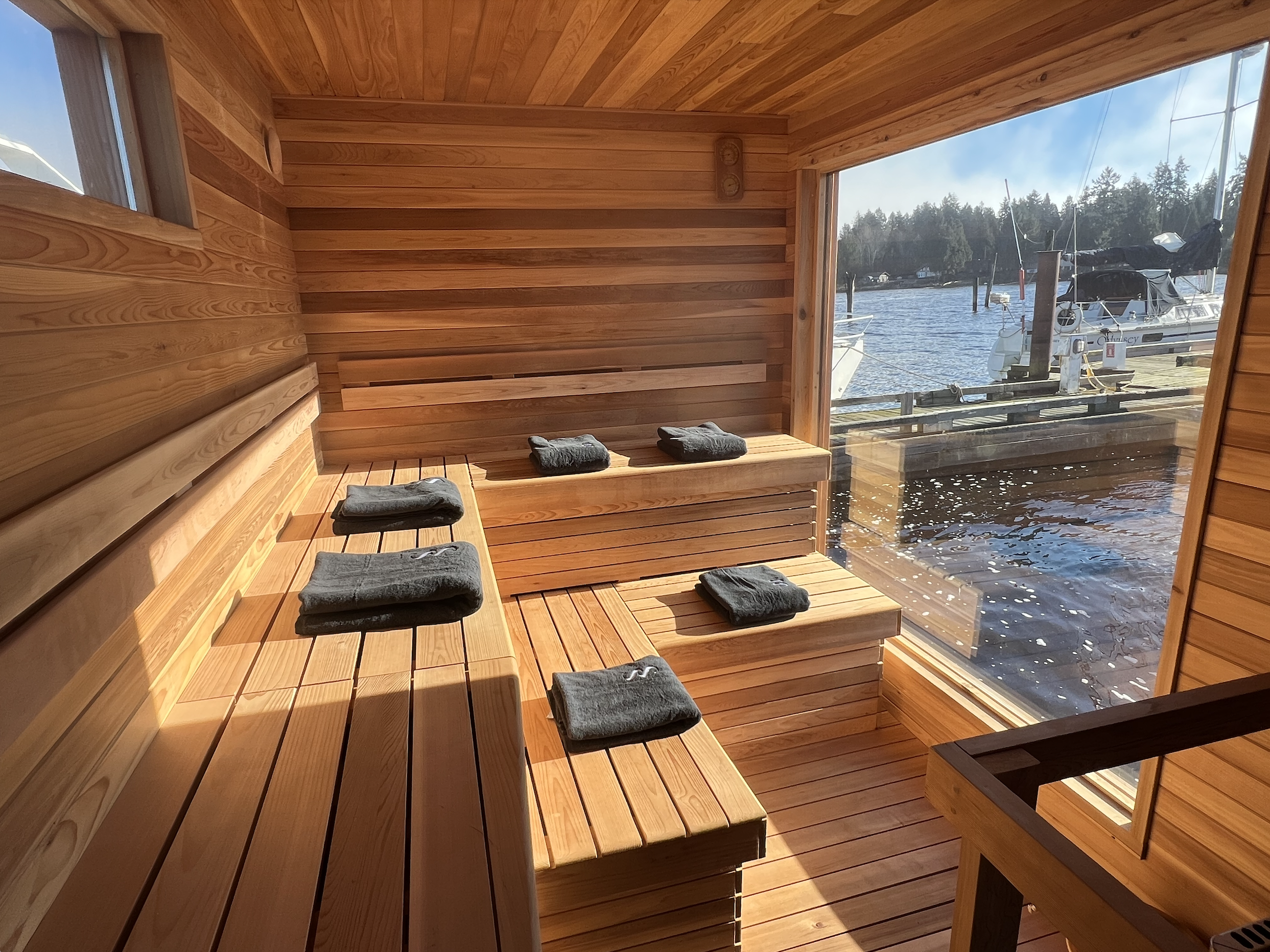 A wooden sauna room with bench seating and towels, overlooking a marina with boats through large glass windows.