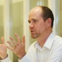 A man with short hair and a goatee, wearing a white shirt, gesturing with his hands while talking or explaining something.