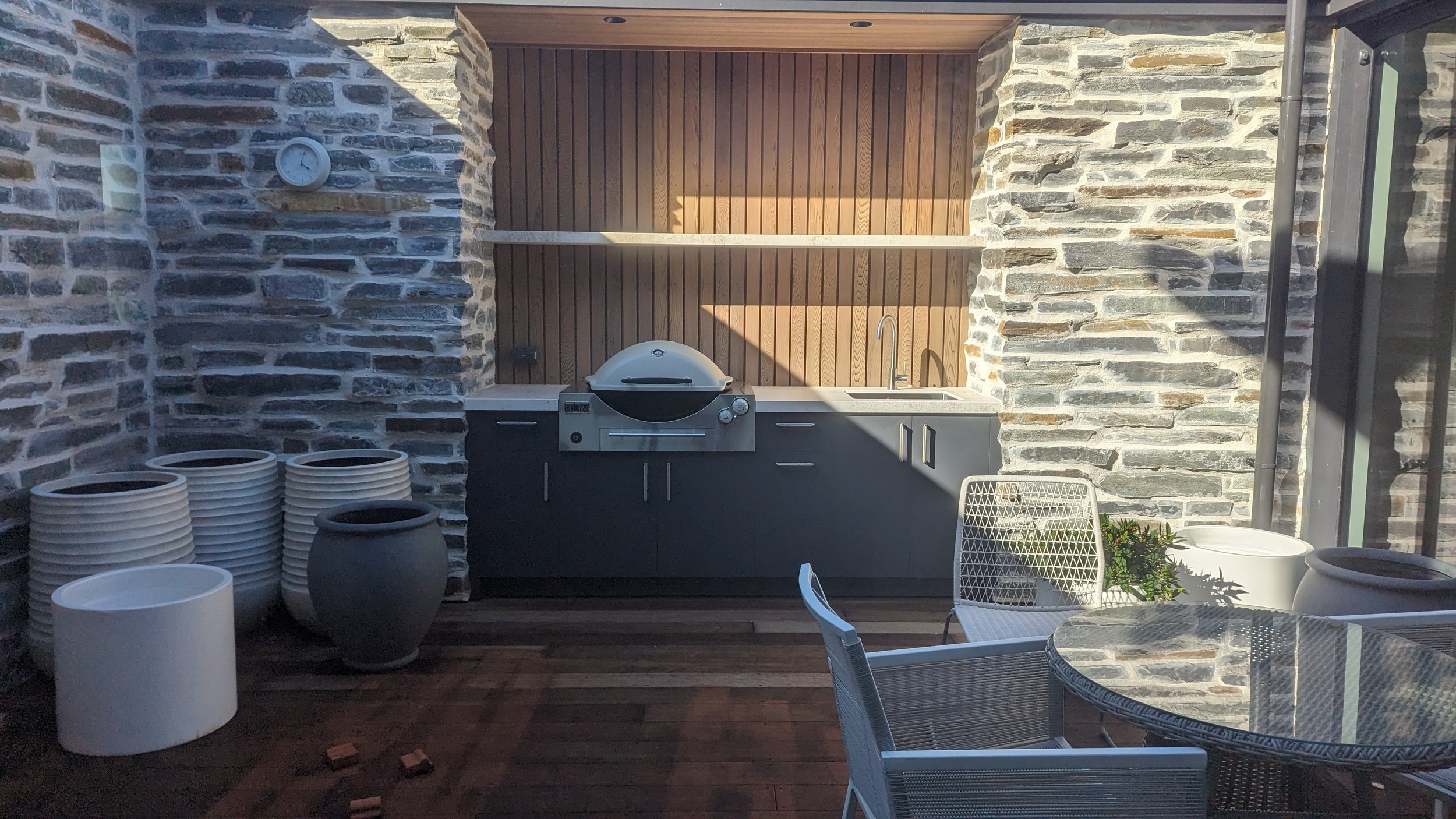 Outdoor kitchen with stone walls, a built-in grill, sink, and countertop, surrounded by white chairs and tables, with large pots and plant decor.