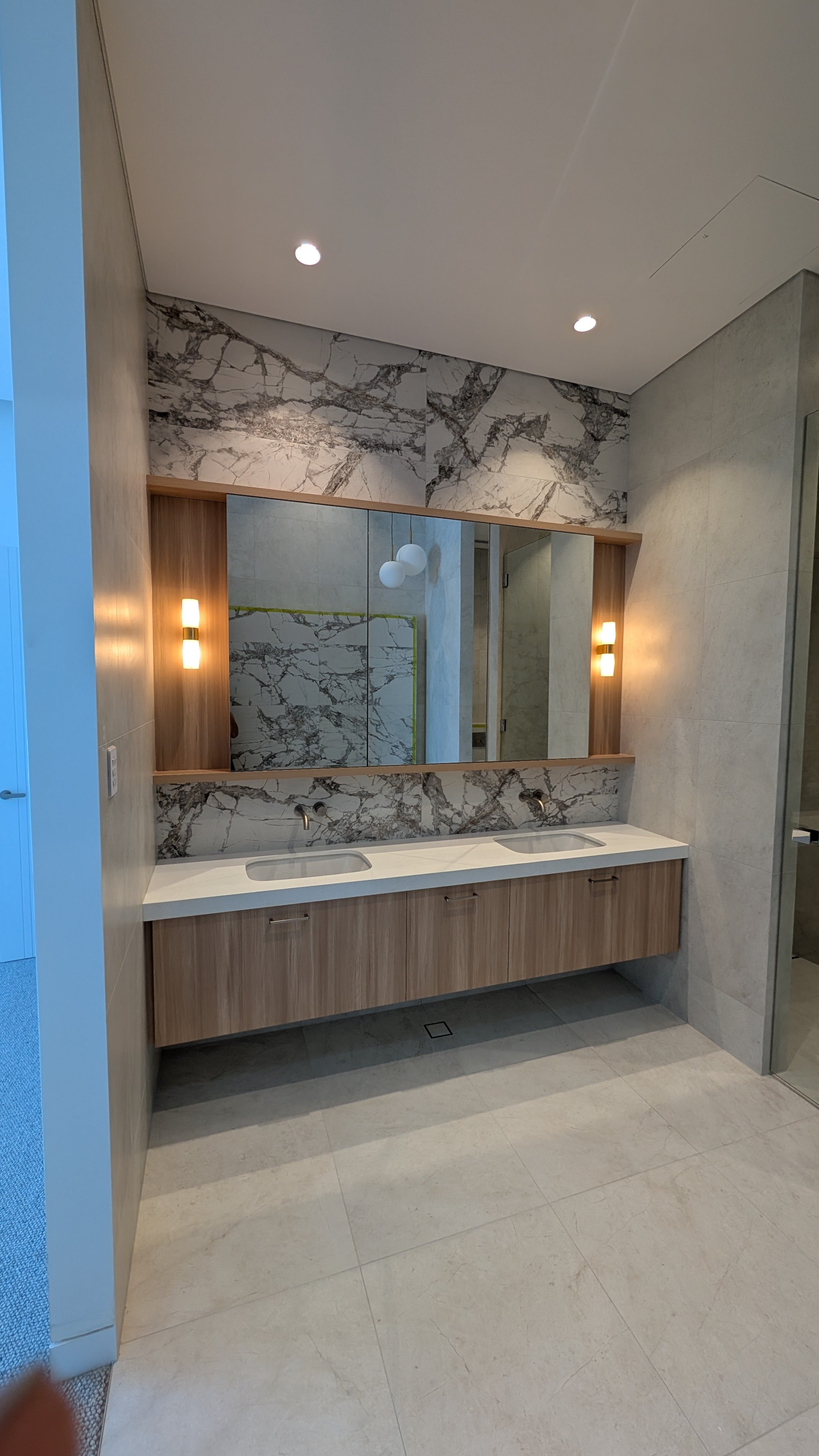 Modern bathroom with double sink vanity, large mirror, marble wall, wooden cabinetry, and minimalist lighting fixtures.
