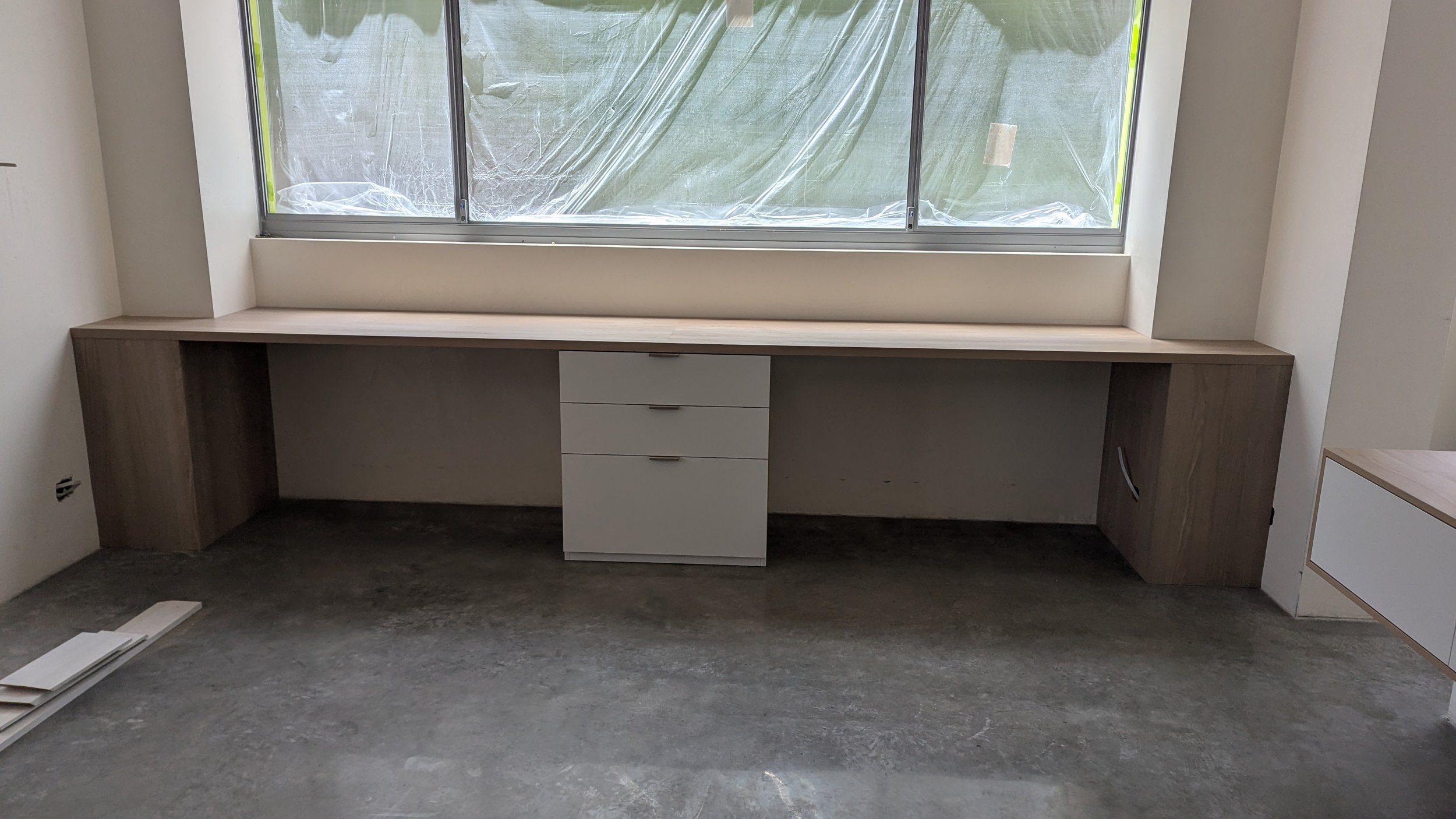 In-progress interior room with a long wooden desk beneath a large window, unclear what the room will be used for.