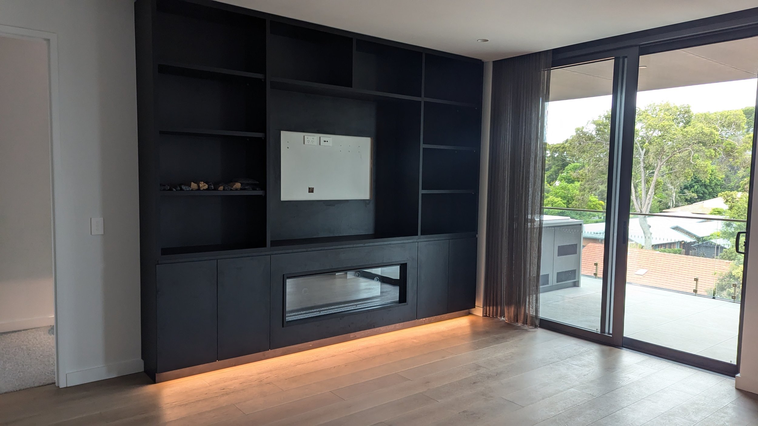 Living room with black built-in shelf and cabinet, white wall, and large glass sliding door leading to a balcony with trees outside.
