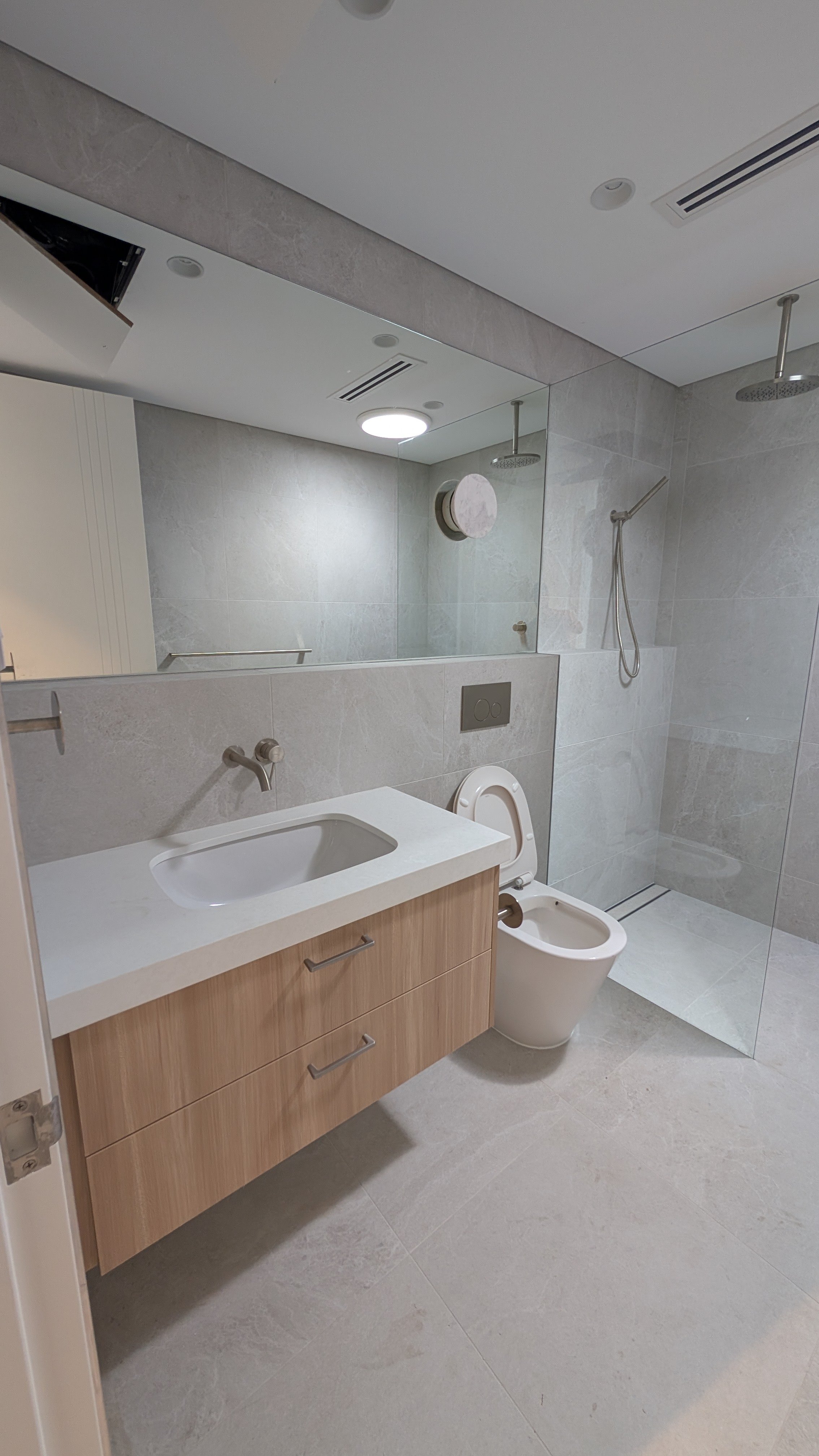 Modern bathroom with a large mirror, a white countertop with a built-in sink, a wooden vanity with two drawers, a wall-mounted toilet, and a walk-in shower with a rainfall showerhead, all with gray tiled walls and floors.
