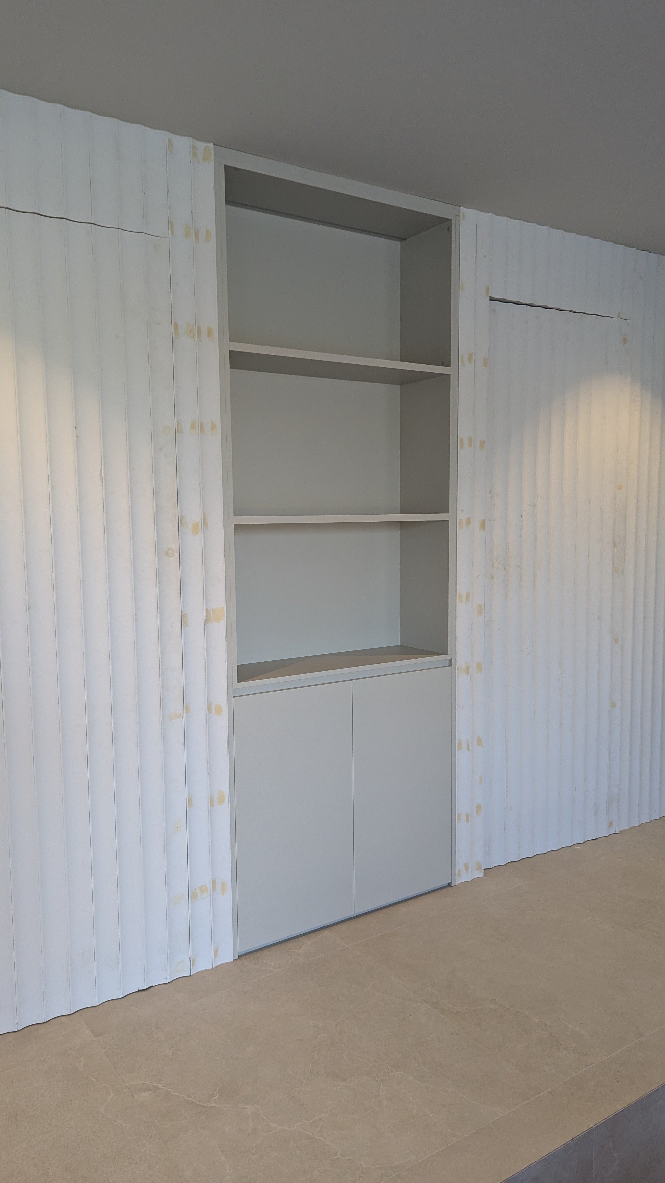 Unfinished built-in white shelving unit with three open shelves and closed cabinets at the bottom, against a wall with textured white and yellow paneling in a room under construction.
