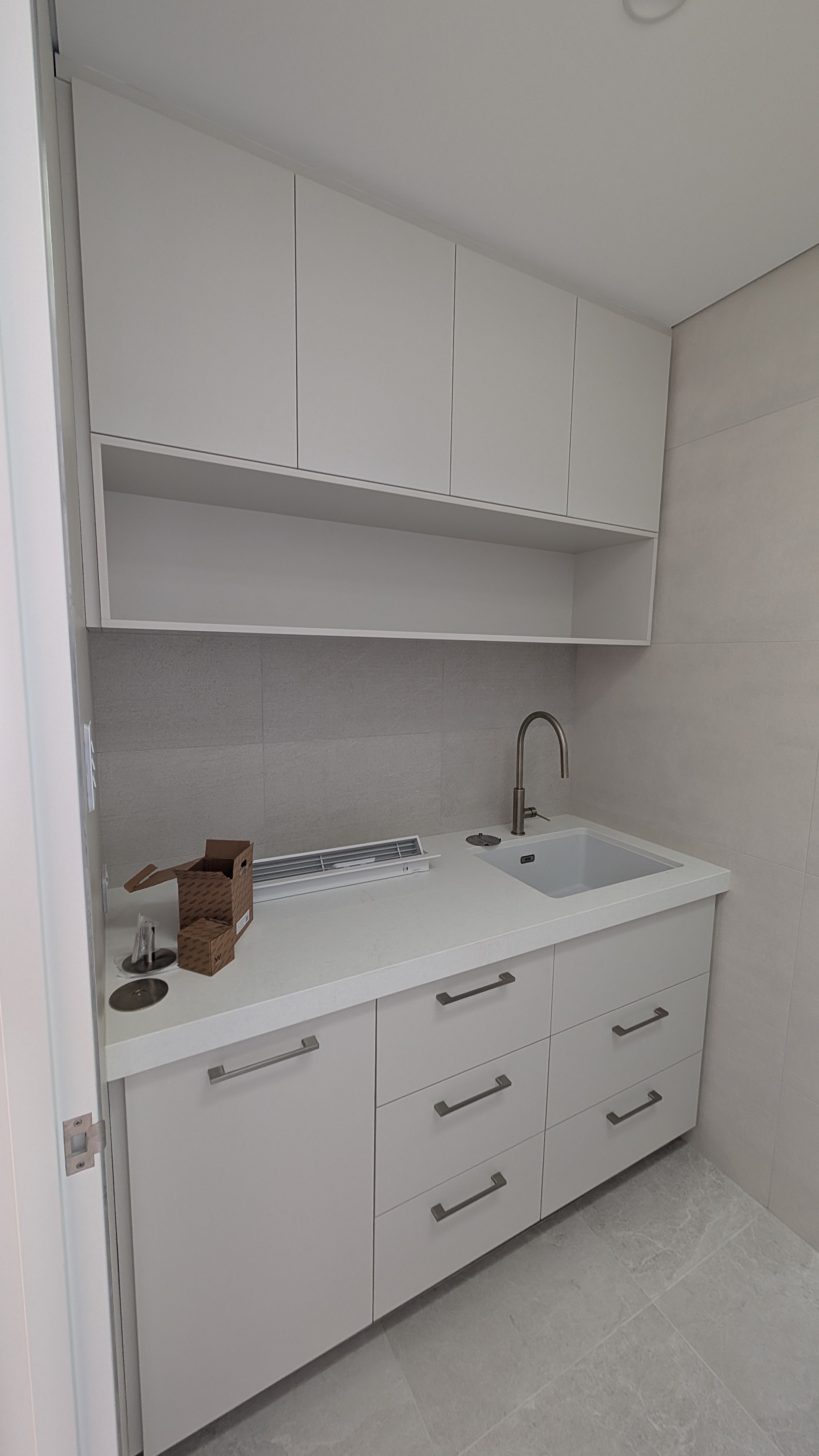 Modern white kitchen with upper cabinets, lower drawers, a sink with a faucet, and some small boxes and a dish drainer on the counter.