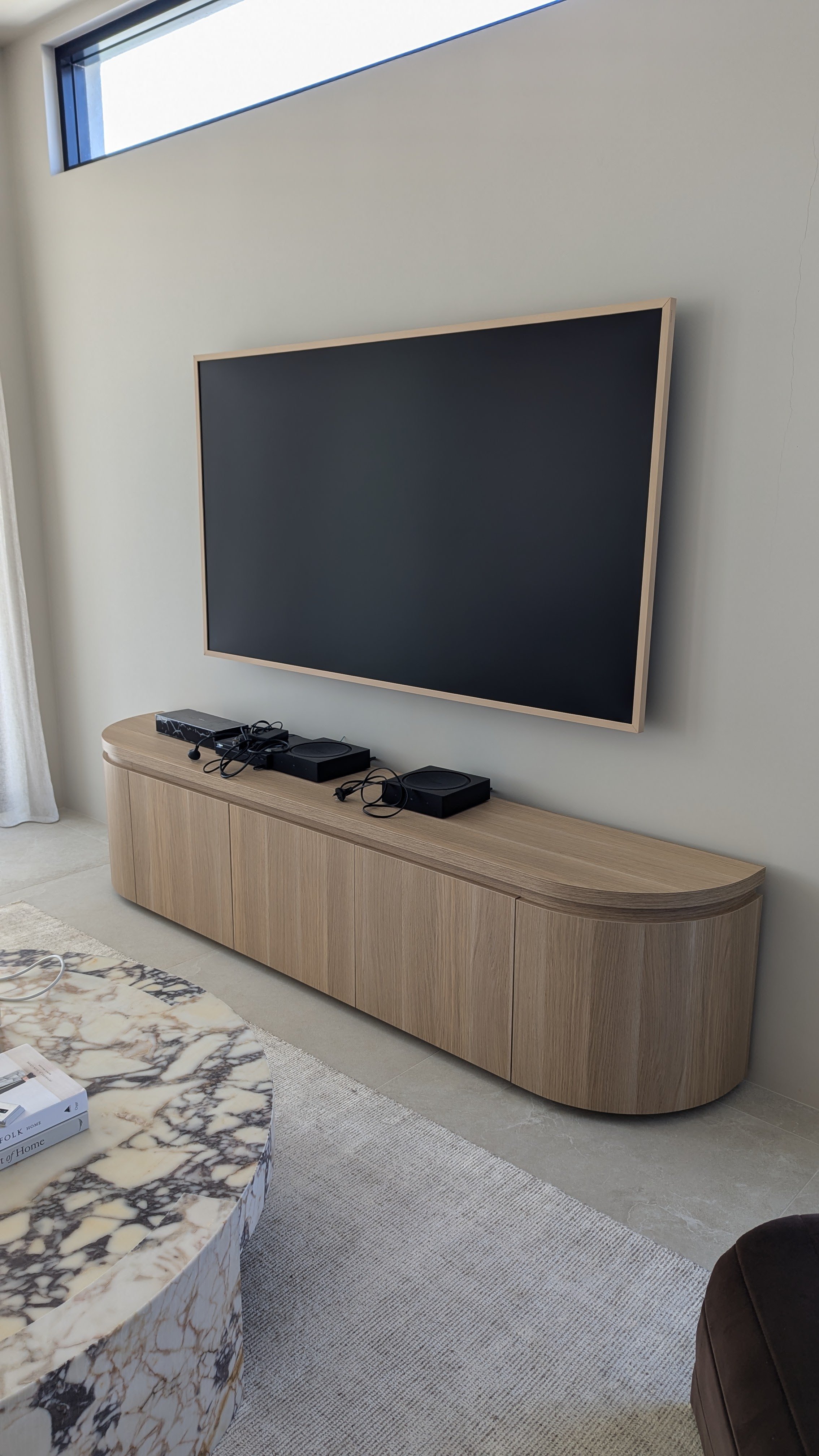 Living room with a large flat-screen TV mounted on the wall above a wooden cabinet. Several electronic devices and cords are on the cabinet. A marble coffee table and a part of a sofa are also visible.