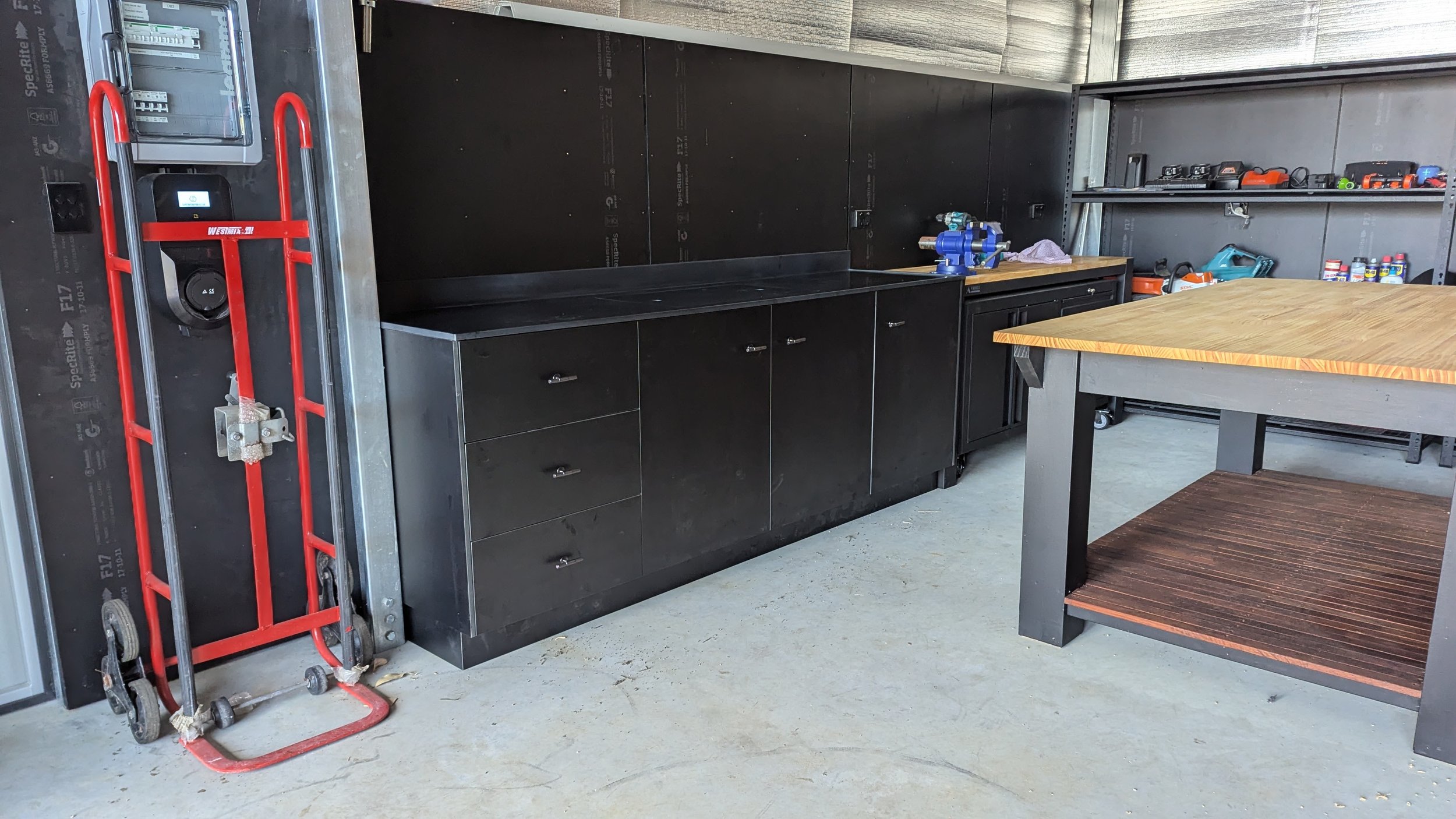 A garage workspace with black cabinets, a wooden workbench, and various tools on shelves, with a red hand truck leaning against the wall on the left side.