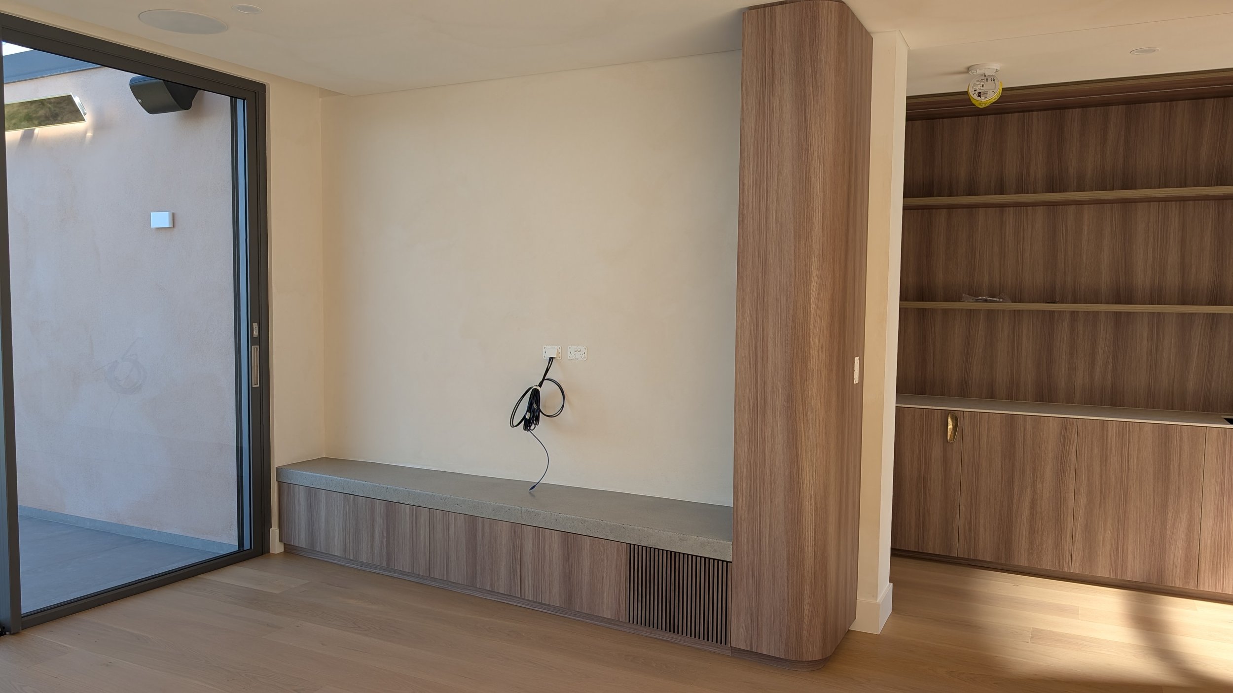 Empty room with a large sliding glass door, a built-in wooden bench under electrical outlets, and a tall wooden bookshelf with sliding doors, in a modern home interior.