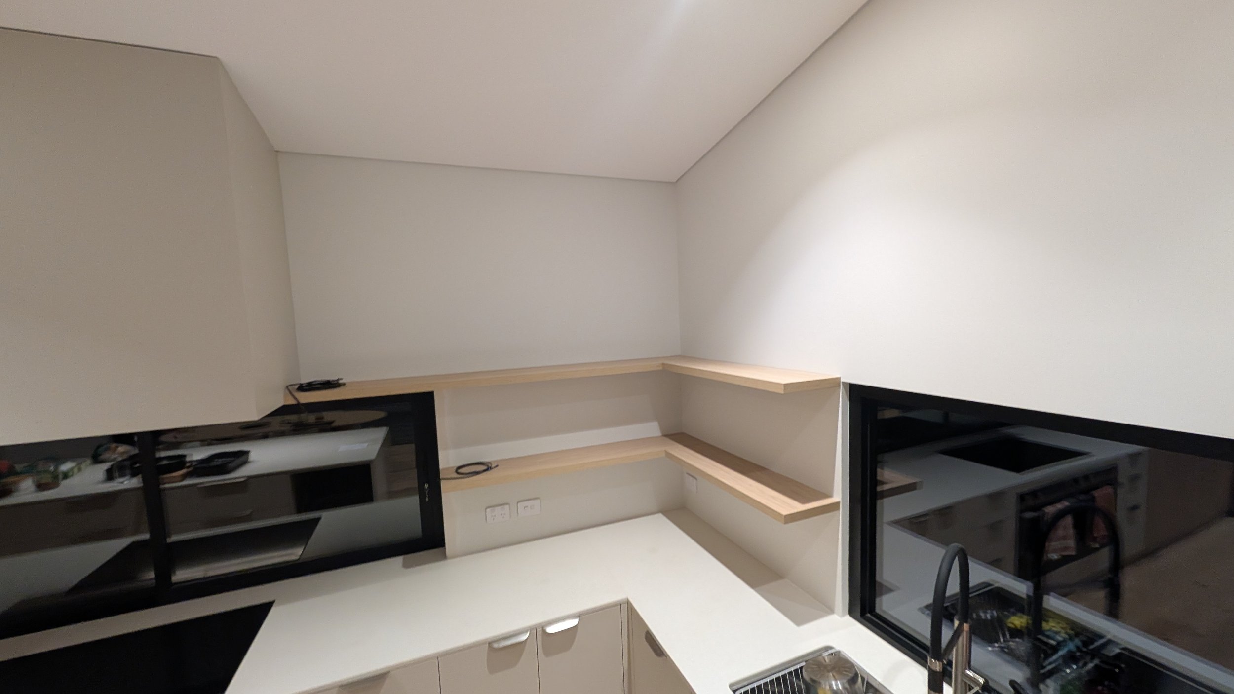 Corner kitchen with white countertops, beige cabinets, and exposed wooden shelves on a wall, with a black-framed window adjacent to a gas stove and sink.