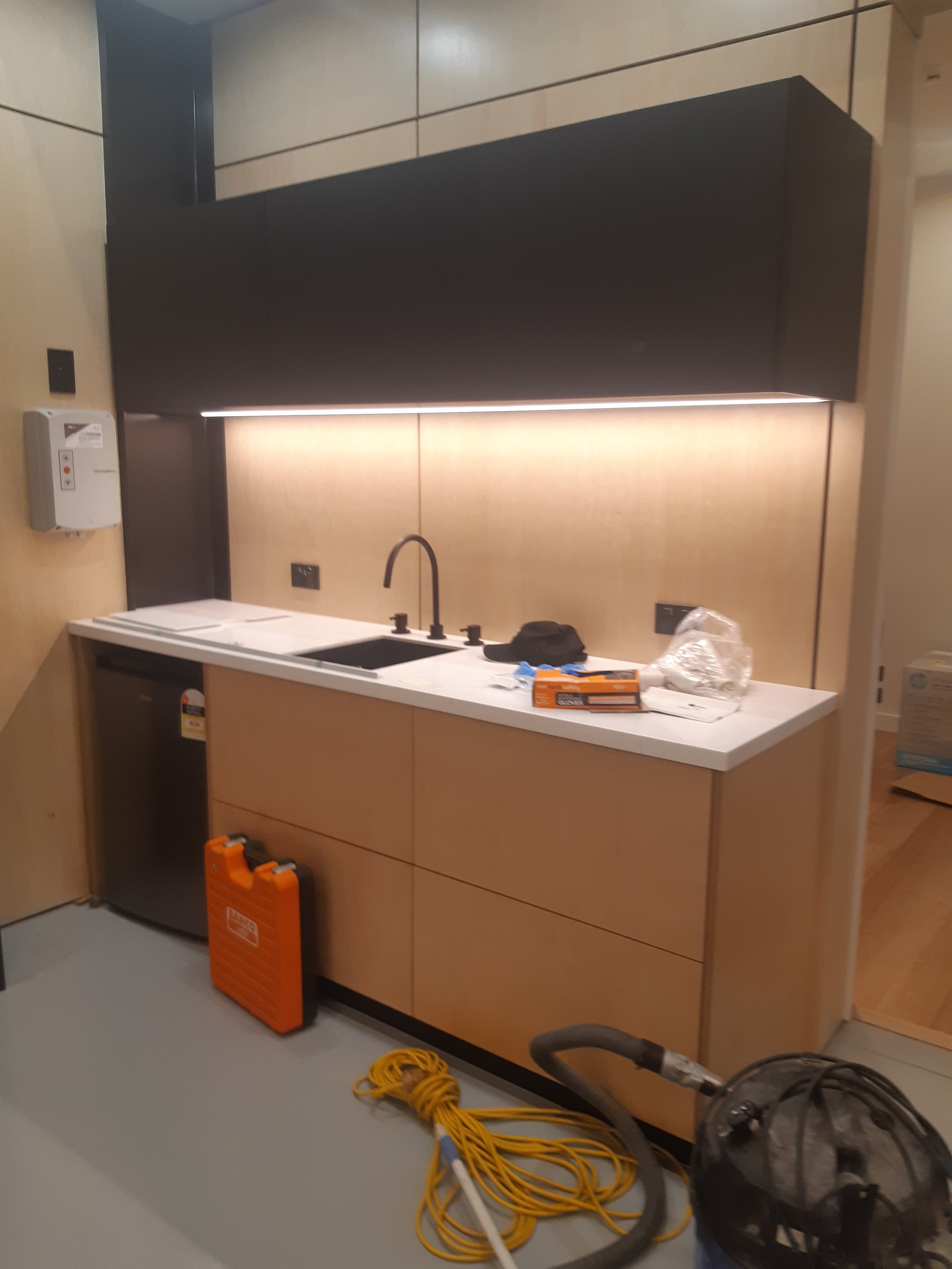 Unfinished kitchenette with a black upper cabinet, a white countertop, and a small sink. There are cleaning tools and construction materials on the counter and floor, indicating ongoing installation or renovation.