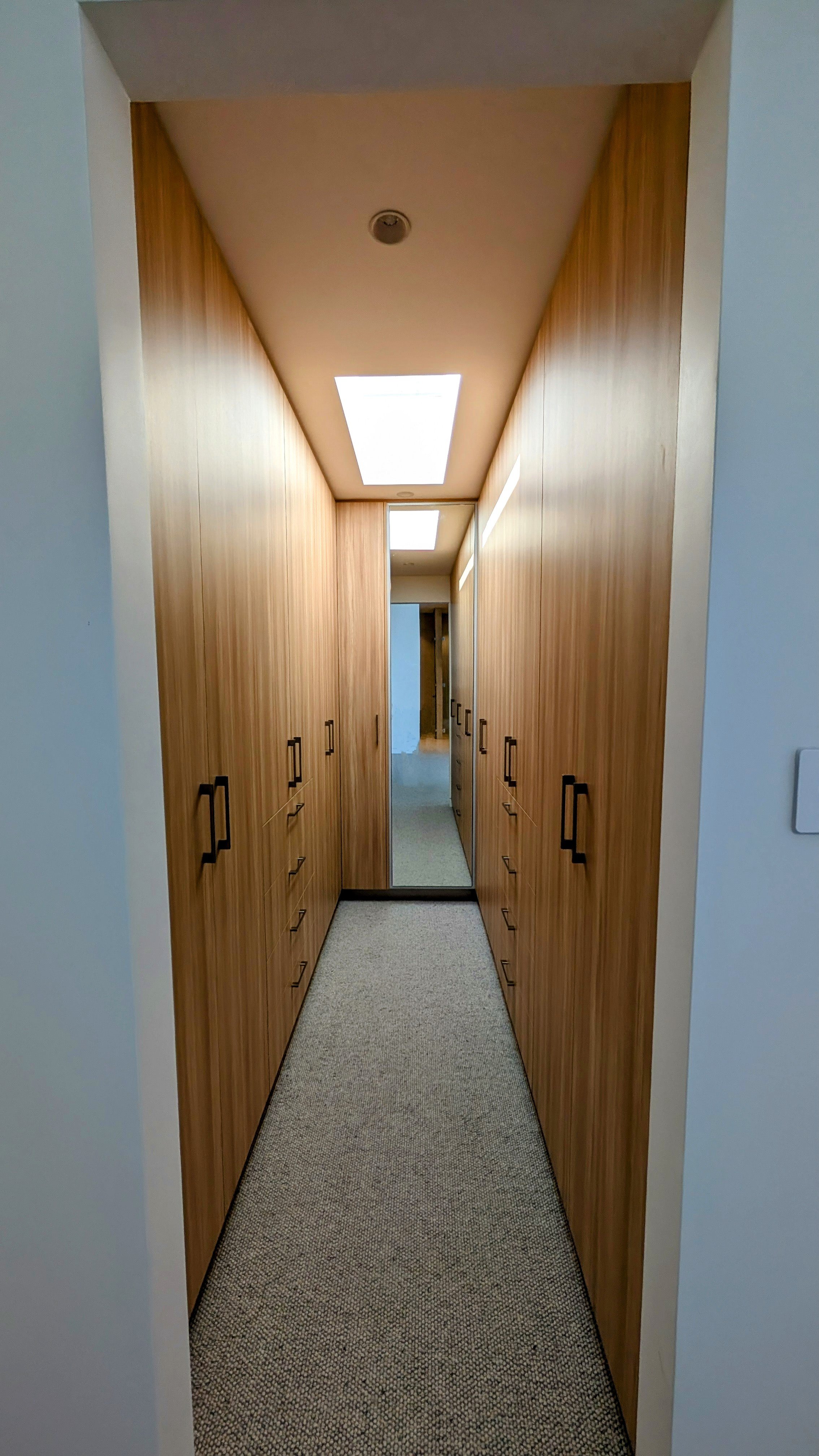A narrow hallway with wooden cabinets on both sides, a mirror at the end, and rectangular ceiling lights.
