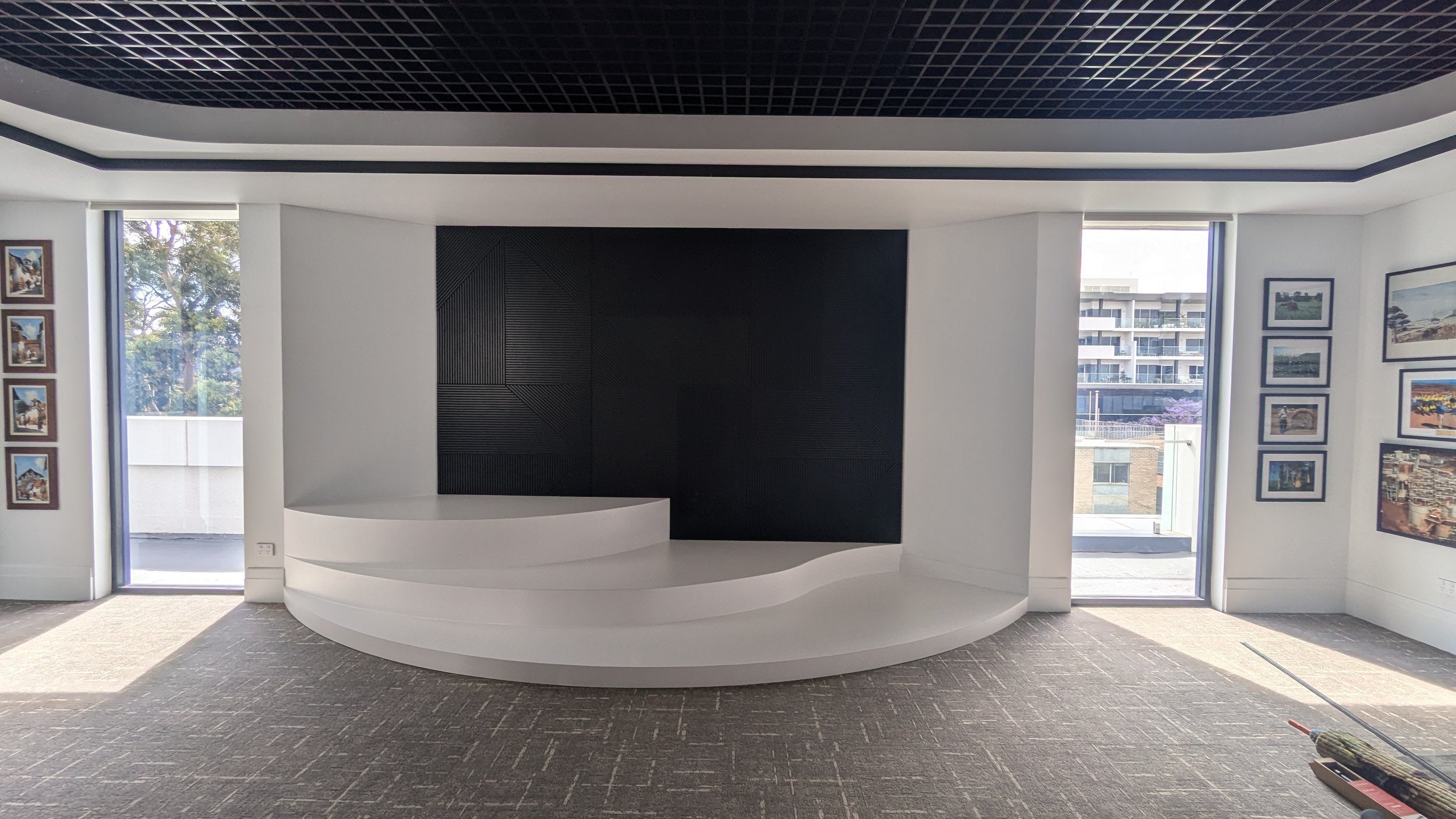 Modern, empty interior room with a white curved platform, black textured wall background, large windows on sides, and framed pictures on white walls.