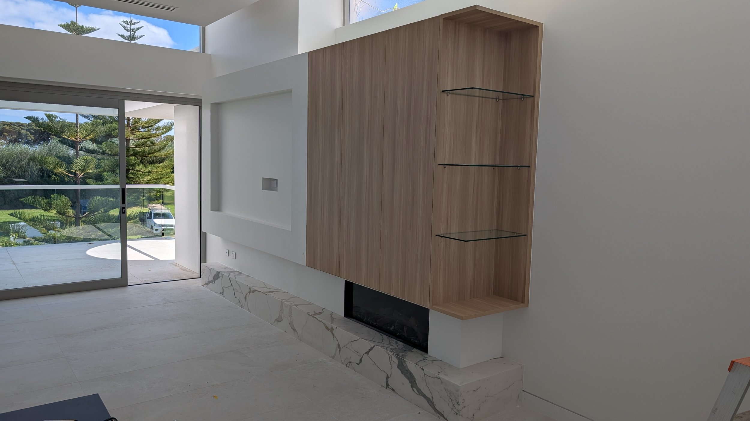 Modern living room with large sliding glass door showing outdoor trees and cars, white walls, polished marble floor, and a built-in wooden shelf with glass panels.