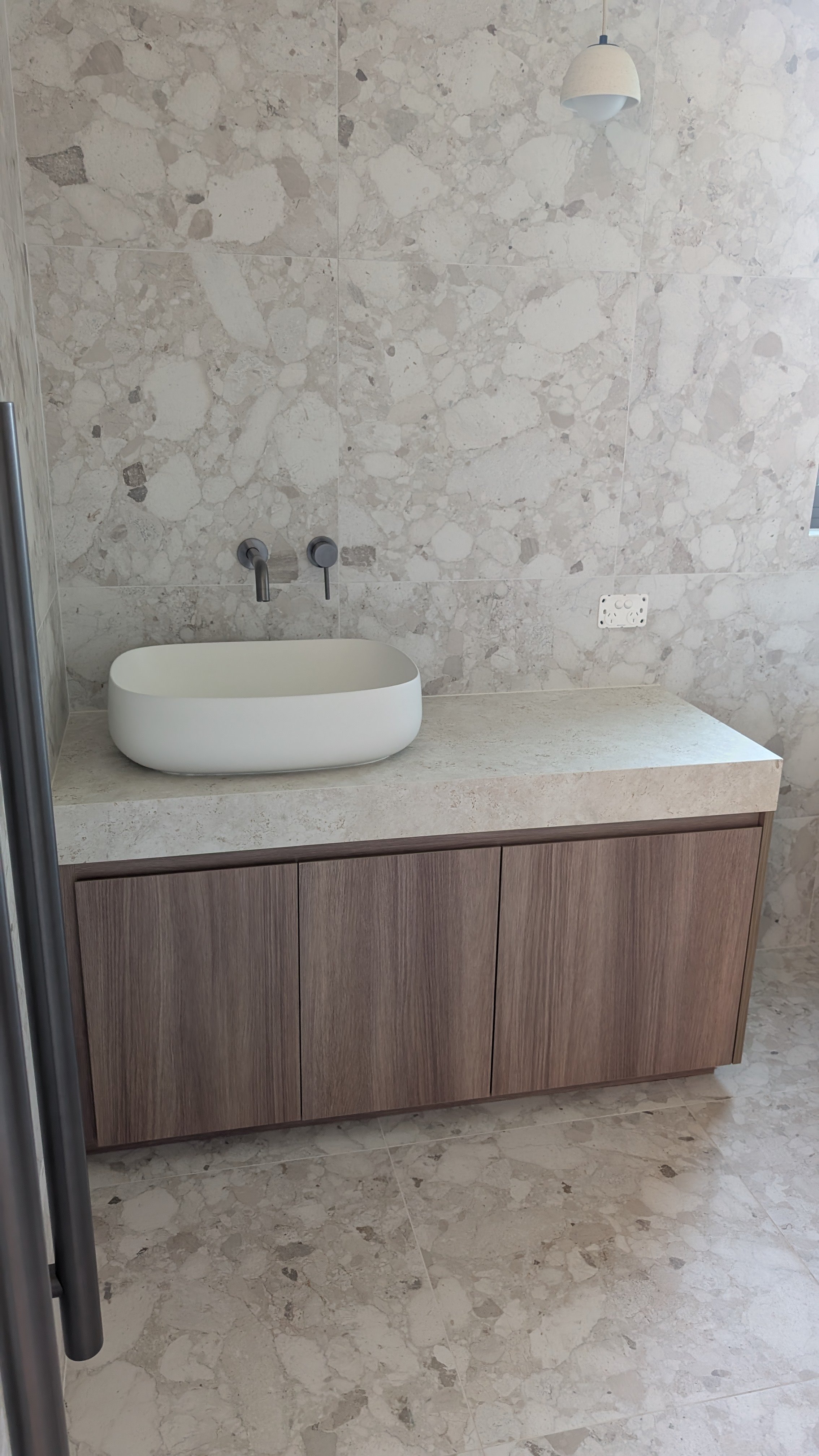 A modern bathroom vanity with a white oval sink, faucet, and seamless wood cabinet below, set against a background of marble wall and floor tiles, with a hanging white ceiling lamp.