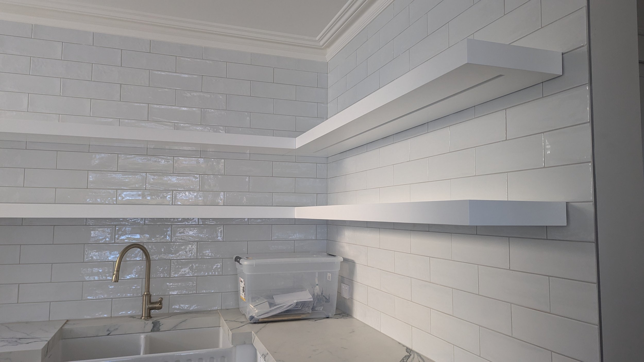 Kitchen with white subway tile backsplash, white floating shelves, marble countertop, and a gold faucet over a white sink, with a plastic storage container on the counter.