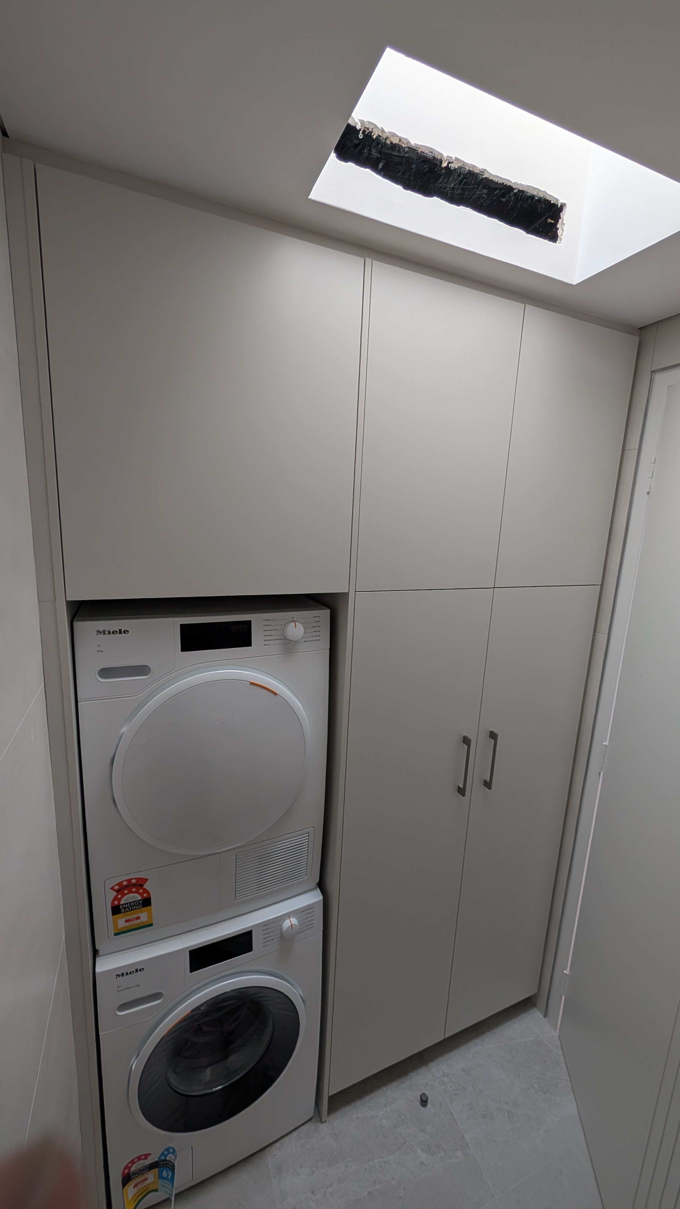 A laundry room with a stacked washer and dryer and closed white cabinets, ceiling with a skylight.