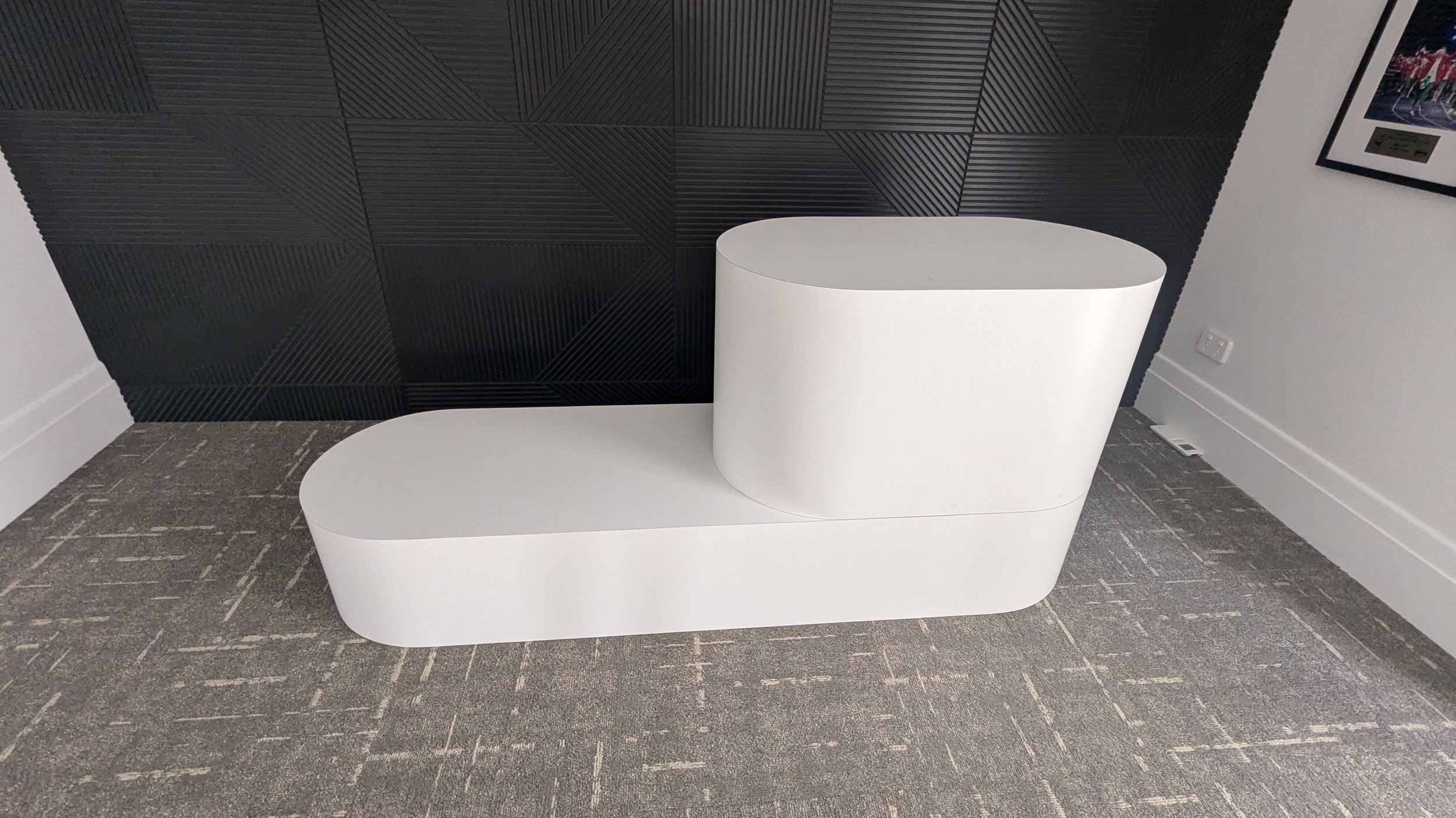 Modern white, curved, two-tiered bench with a circular top, placed against a black textured wall on a carpeted floor.