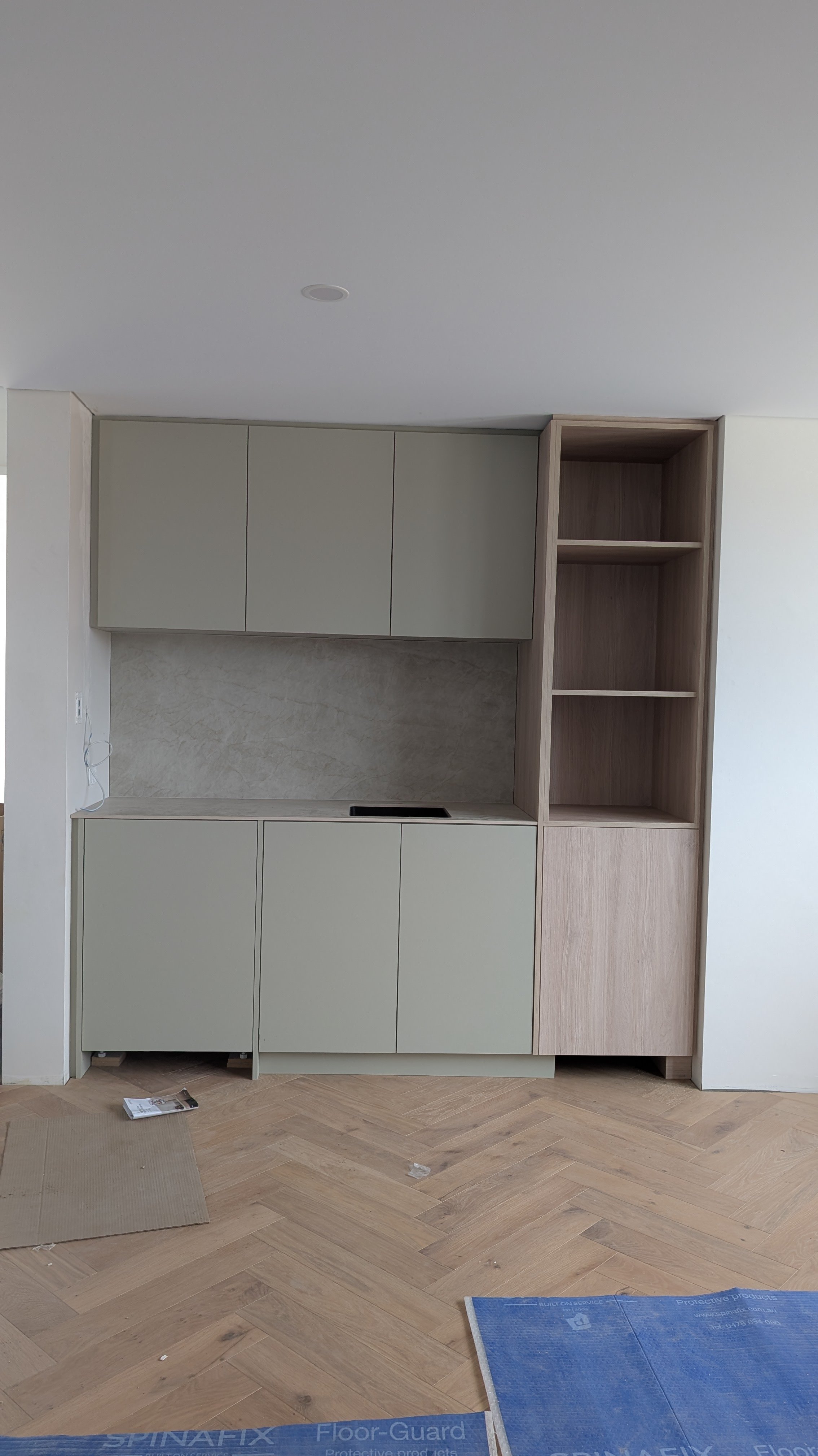 Modern kitchen with beige cabinets, a small stovetop, and open wooden shelving on the right, with unfinished flooring and construction materials on the floor.