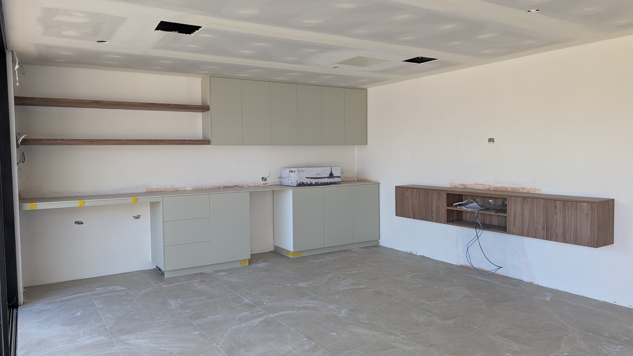 Interior of a kitchen under construction with light-colored cabinets, open shelving, and an uninstalled wall-mounted cabinet, with construction materials and wires visible.