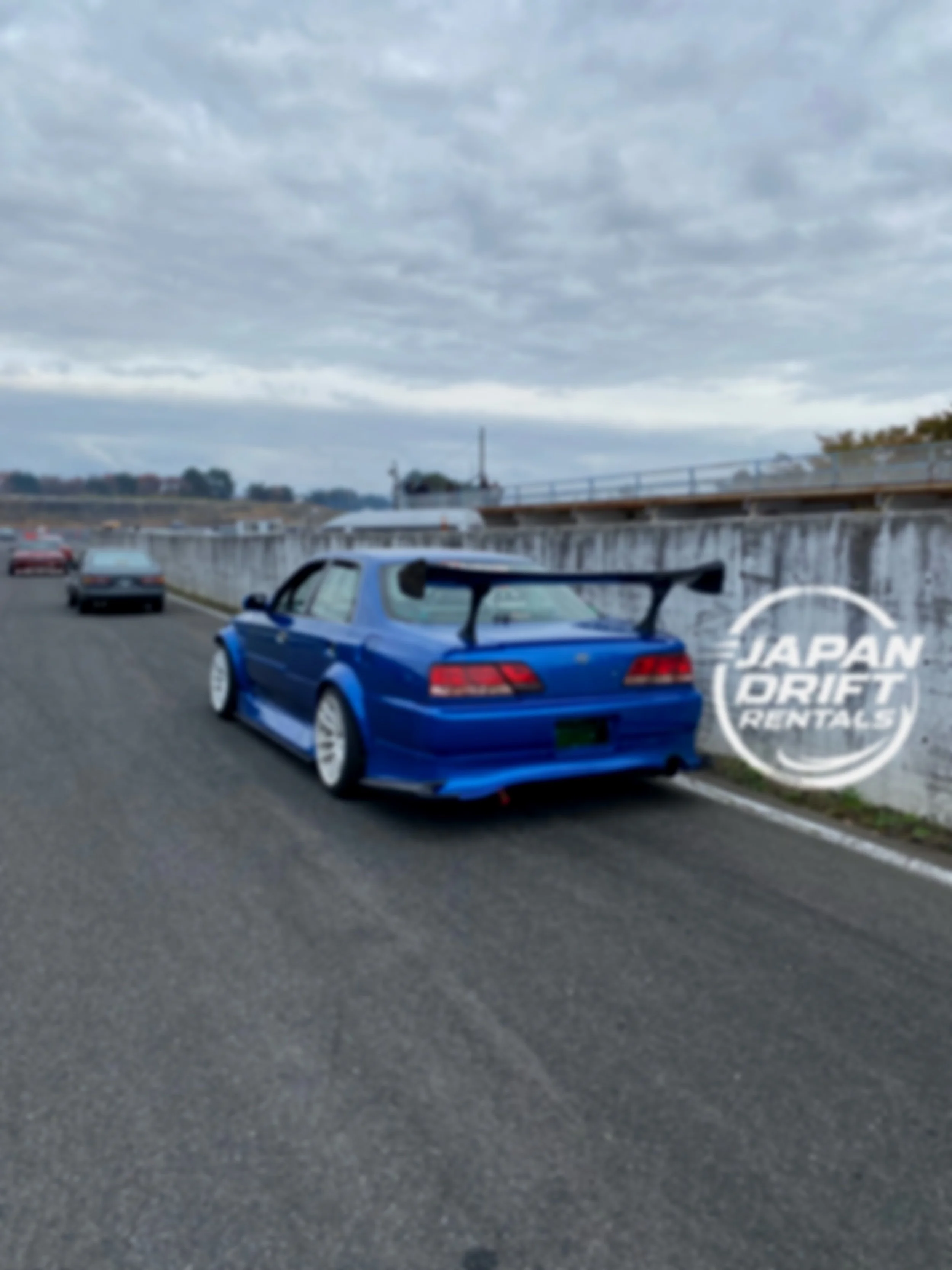 Blue sports car with a large rear wing parked on the side of a roadway, with other vehicles and a concrete wall, under a cloudy sky.