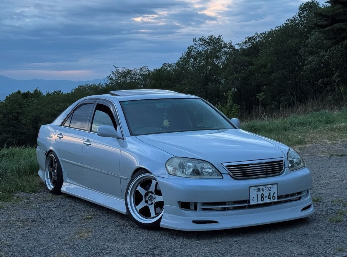 A white modified sedan car jzx110 parked on a gravel area with green grass and trees in the background during dusk or dawn.