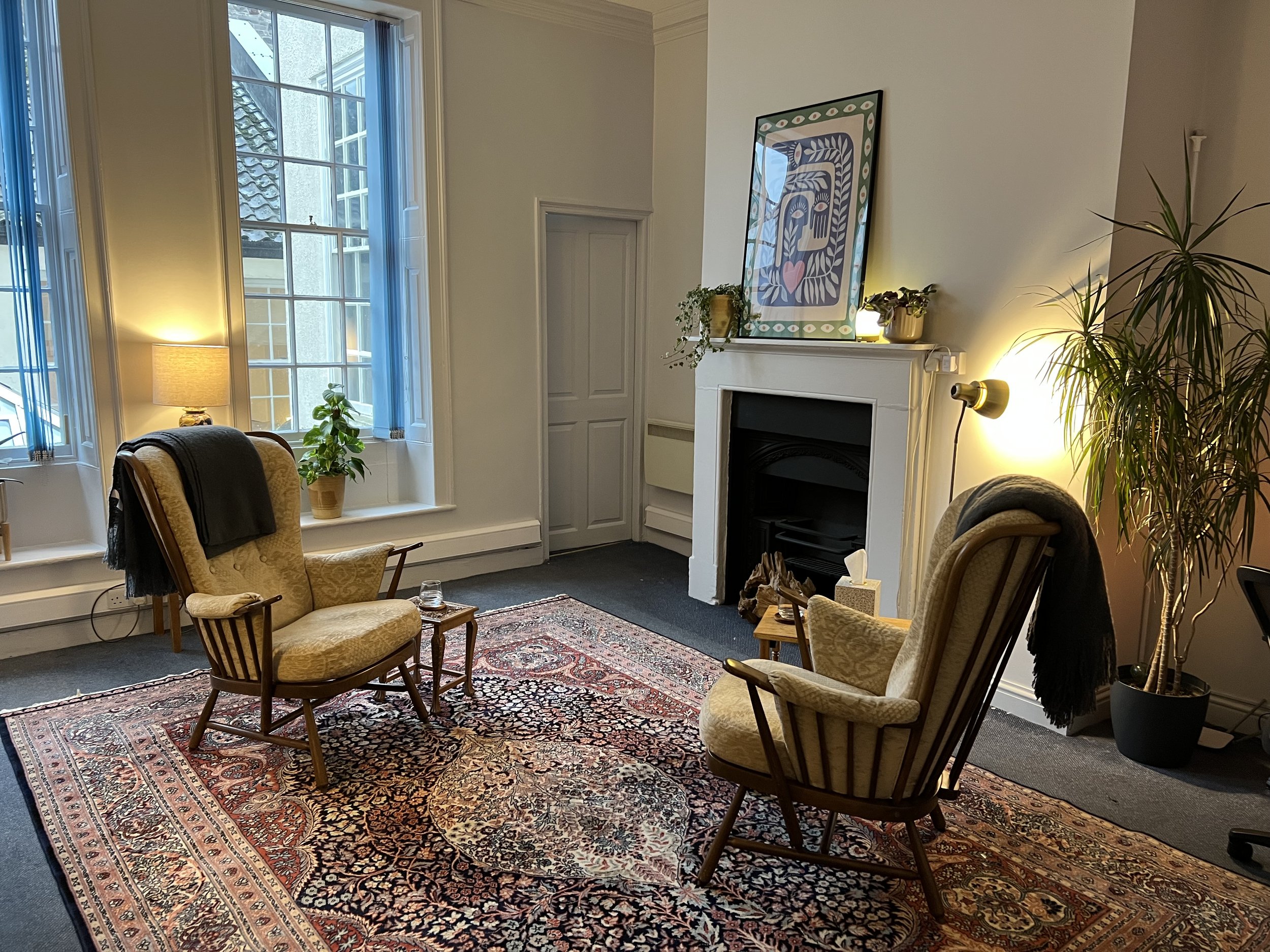 A cozy living room with two yellow upholstered armchairs with wooden armrests on a patterned rug, a small wooden side table, a large window with natural light, a tall potted plant, a white fireplace with decorative items on the mantel, and framed wall art above it.