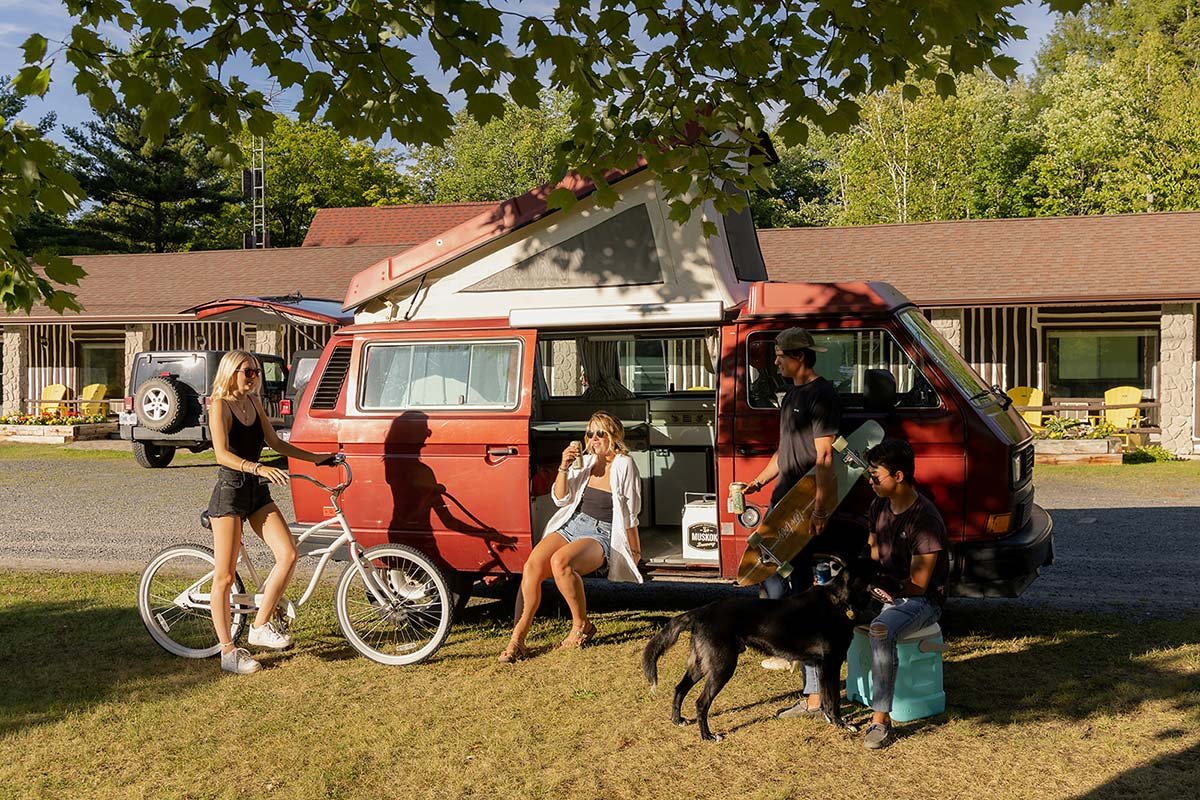 Group of people with a red camper van in a sunny outdoor setting, includes a bicycle, a dog, and a skateboard.