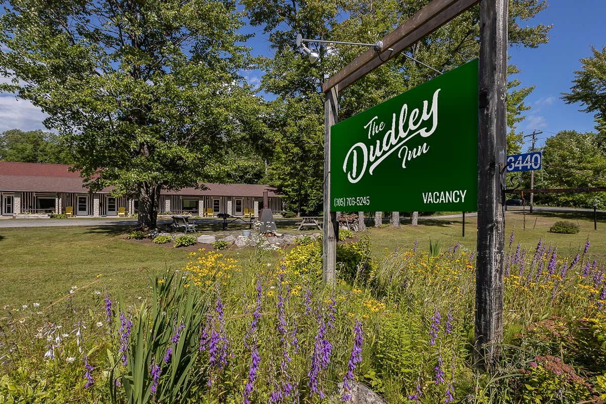 A green sign for The Dudley Inn with the word 'Vacancy' is in the foreground against a backdrop of purple and yellow flowers. A long, single-story building with several windows and a brown roof is in the background, surrounded by trees and grass.