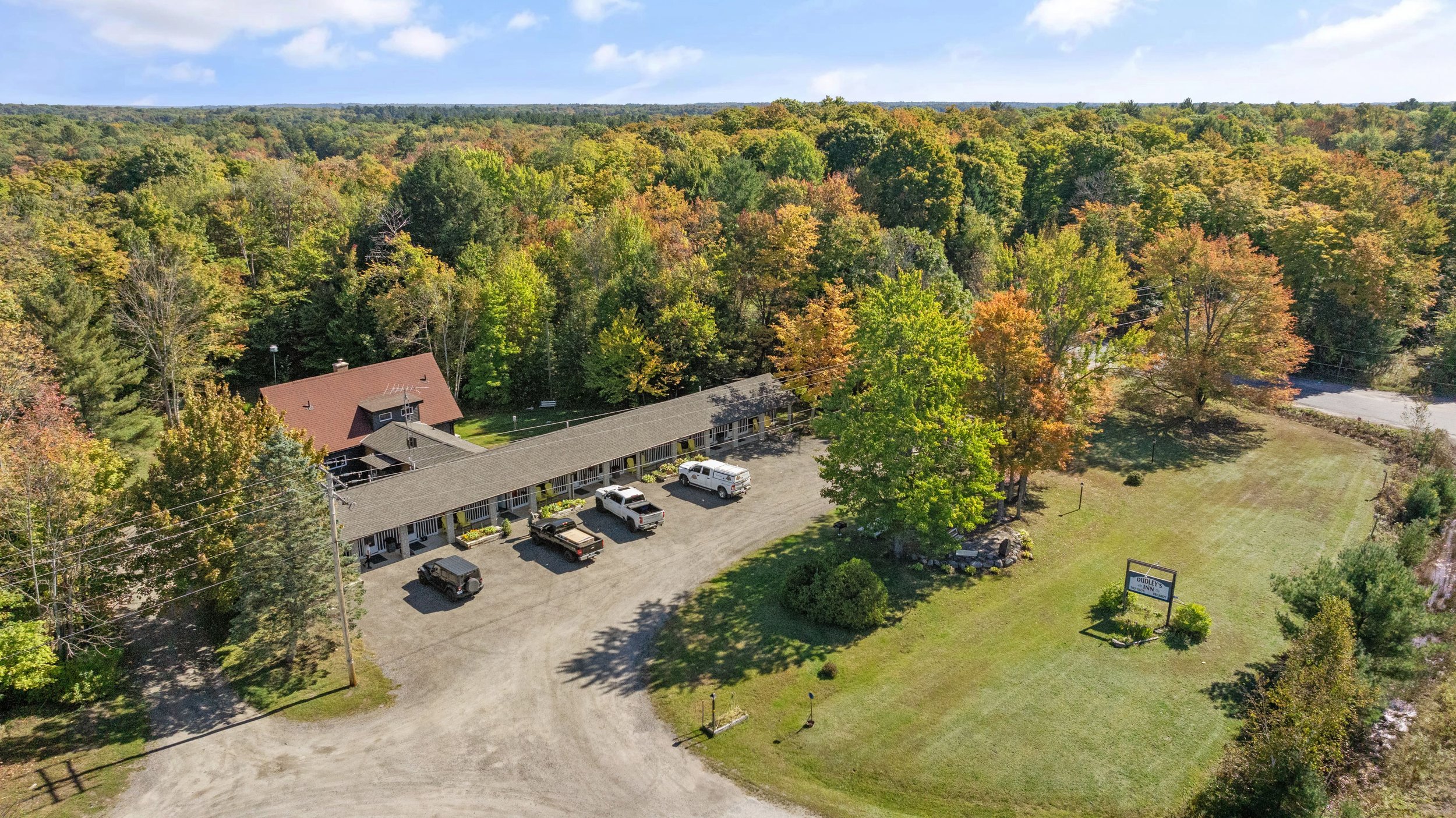 Aerial view of a small rural motel surrounded by lush greenery and trees showing autumn colors. Several vehicles are parked in front of the single-story building. A sign is visible next to a grassy area, and a dense forest is in the background.