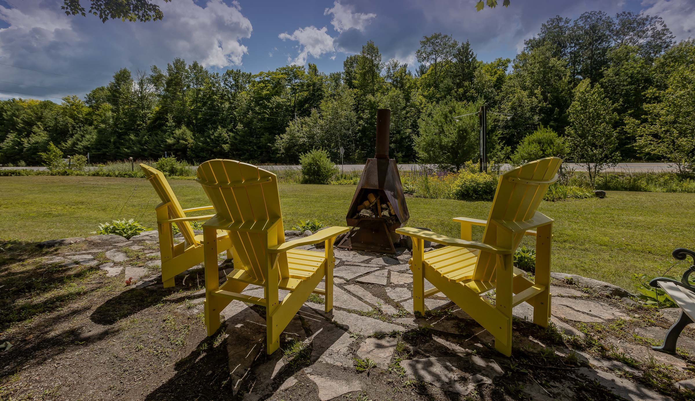 Three yellow Adirondack chairs around a metal fire pit on a stone patio, surrounded by grass and trees in a sunny outdoor setting.