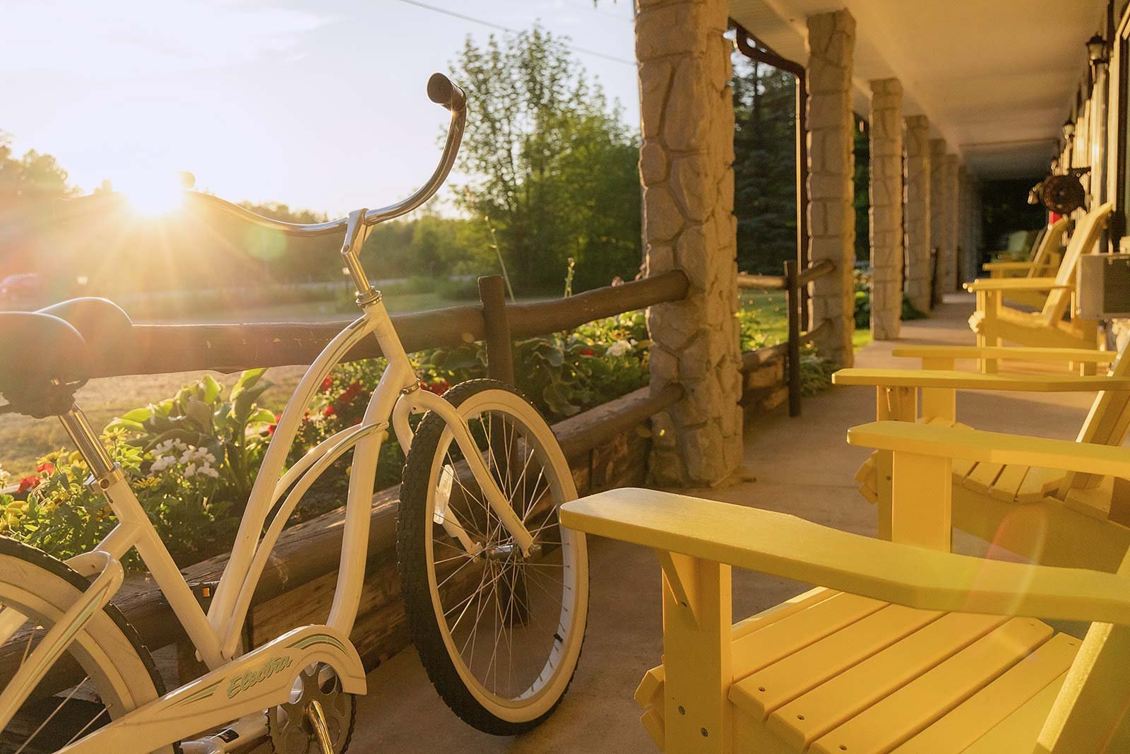 Sunlit porch scene with a white bicycle, Adirondack chairs, and blooming flowers.