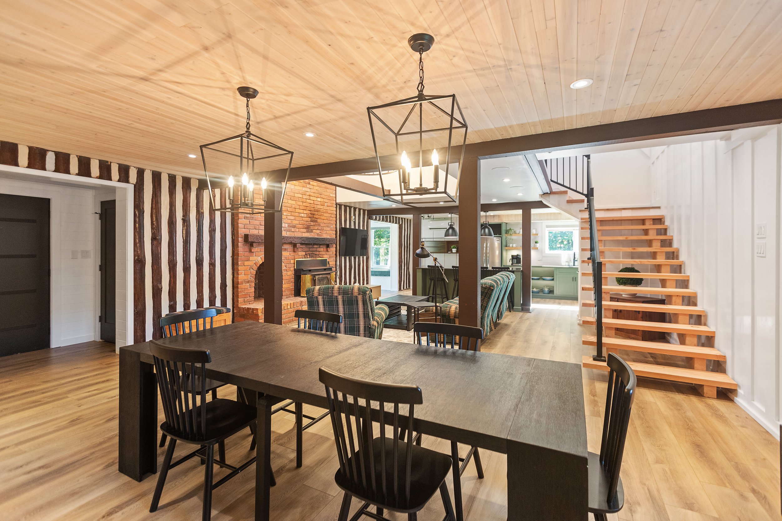 Open-concept living space featuring a wooden dining table with black chairs, modern chandeliers, a brick fireplace, plaid armchairs, and a staircase leading upstairs. The room has a mix of wood and neutral tones with a contemporary design.