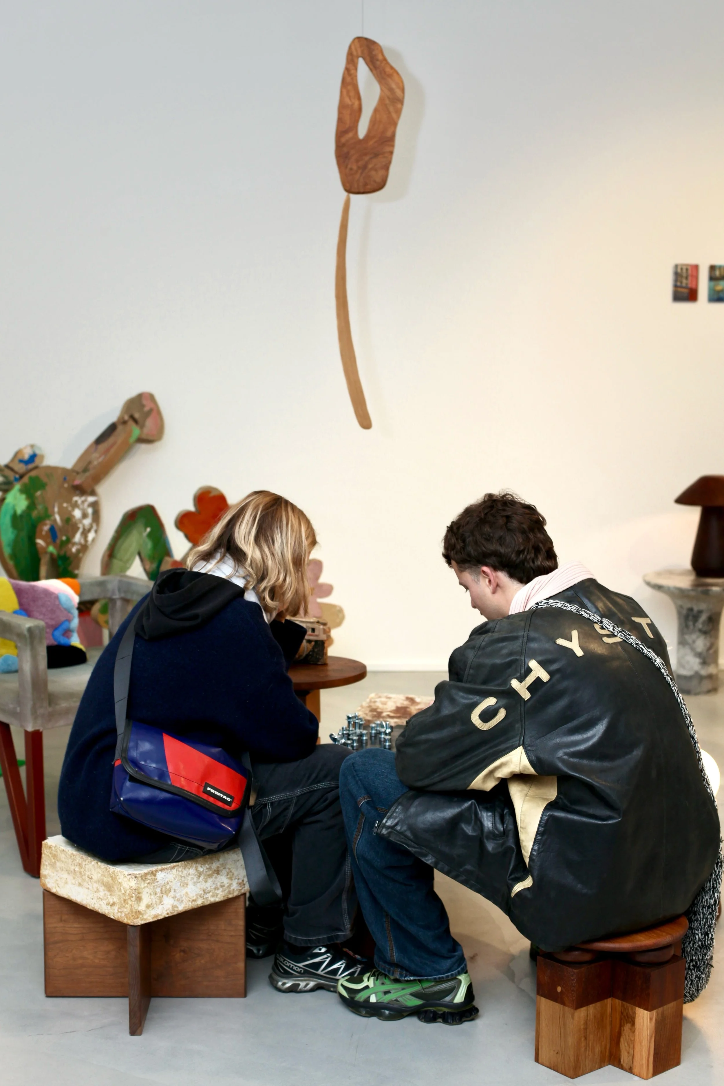 Two young boys sitting closely together on small stools, playing chess in an art gallery or museum, surrounded by artwork and sculptures on the wall.